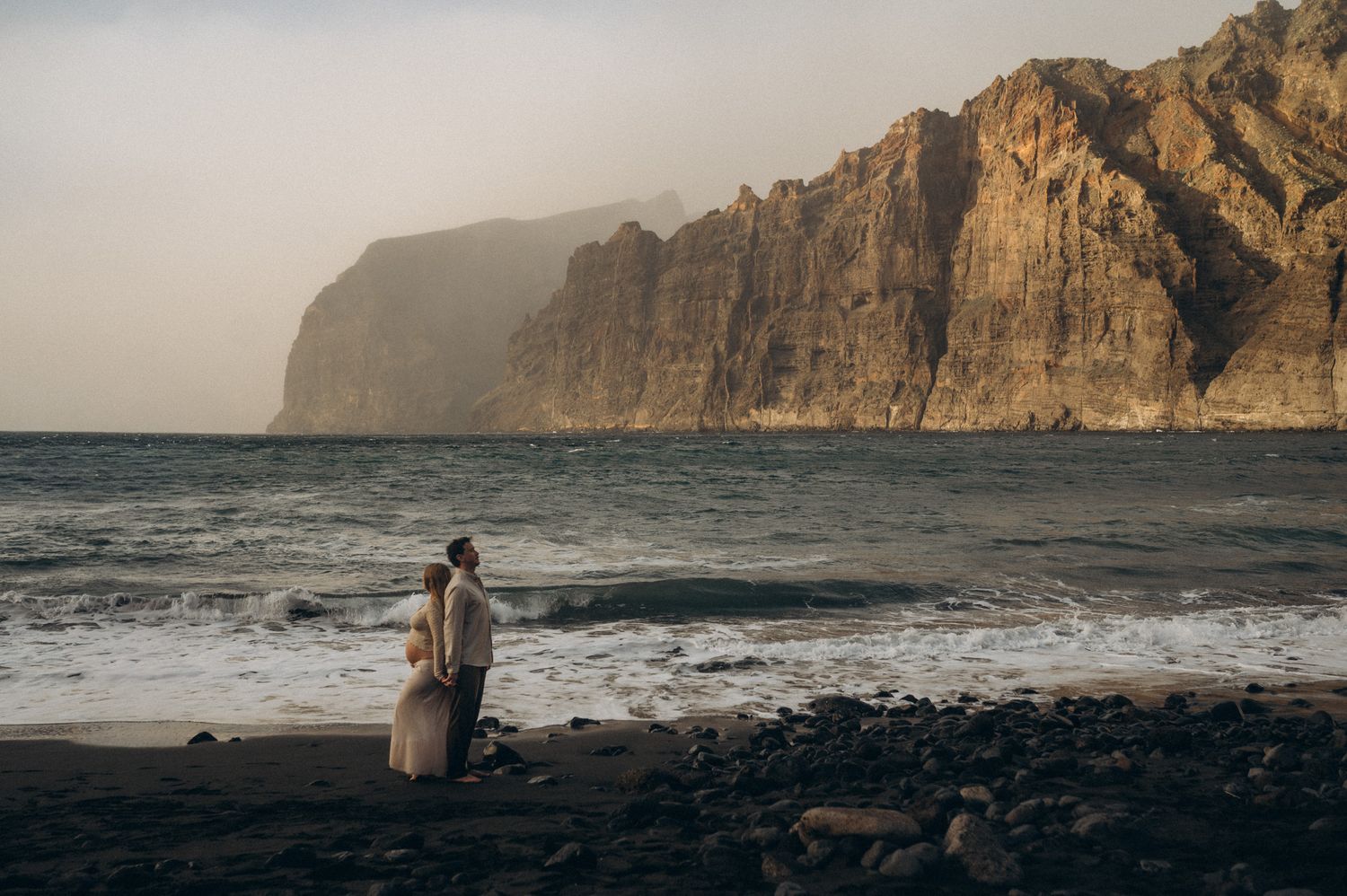 Couple standing on rocky beach with dramatic cliffs and ocean waves at sunset in Tenerife, Spain.