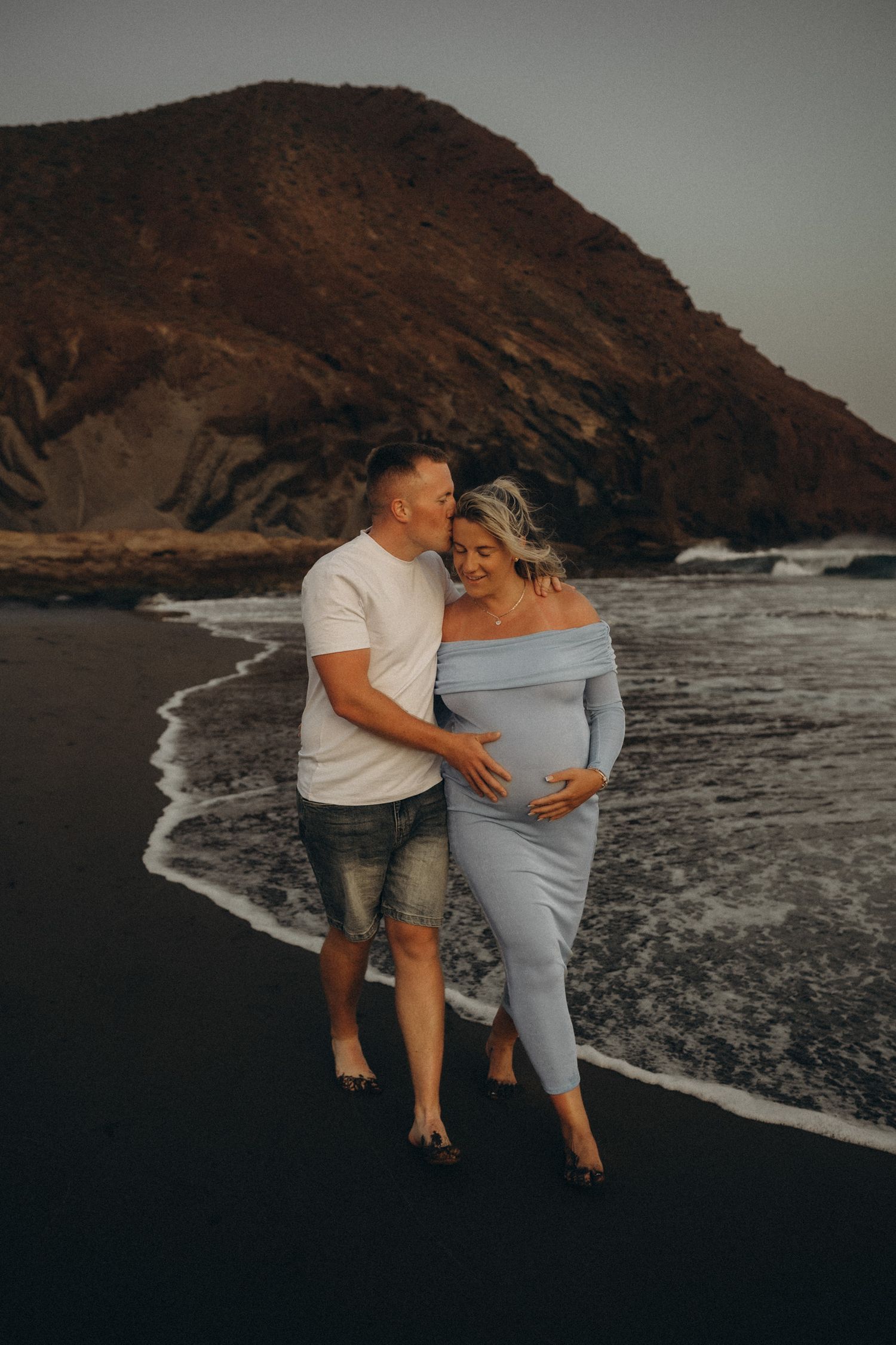 Couple sharing a tender maternity moment on a black sand beach with dramatic cliffs and ocean waves behind them.