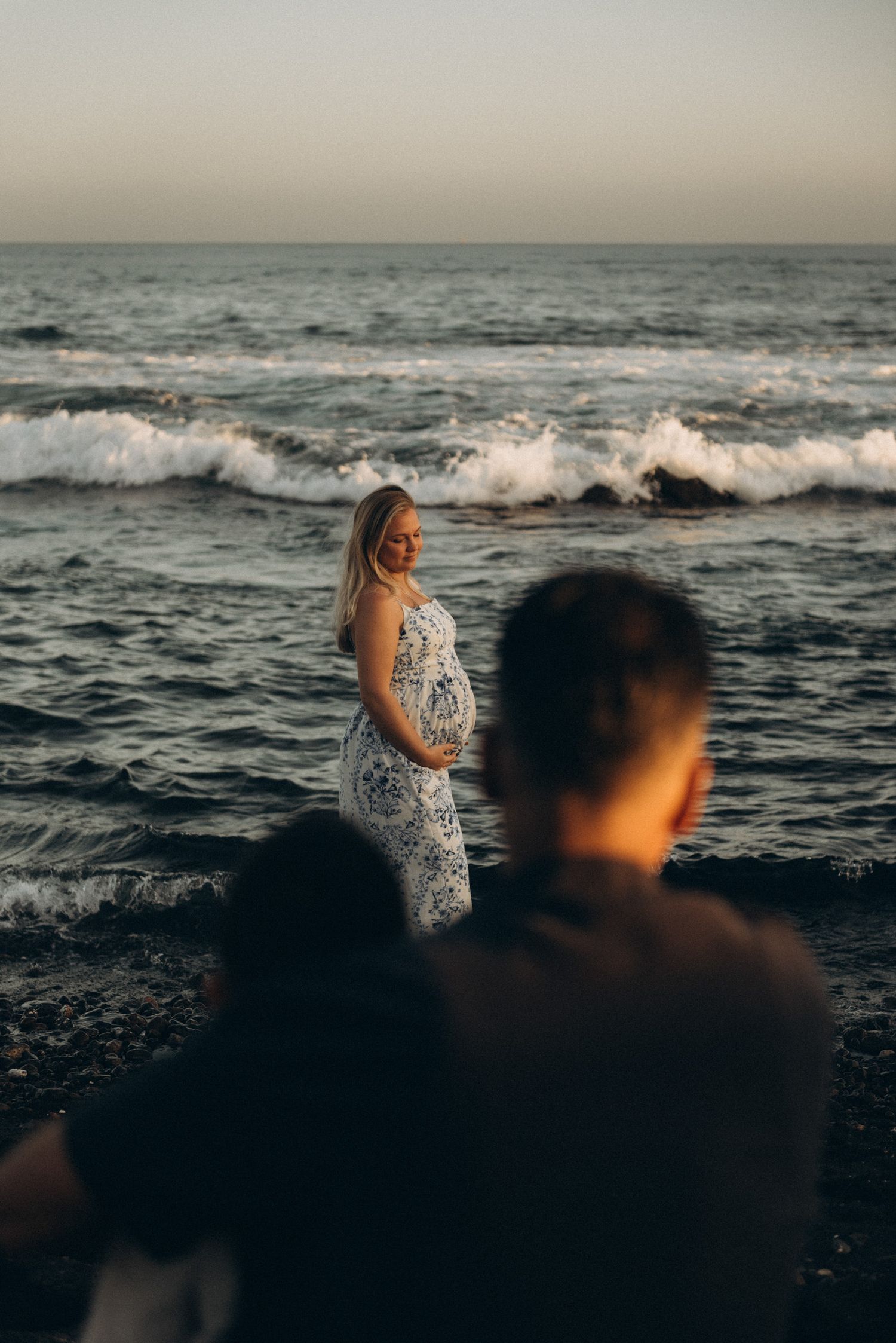 Pregnant woman in floral dress stands by ocean waves at sunset while partner watches nearby.