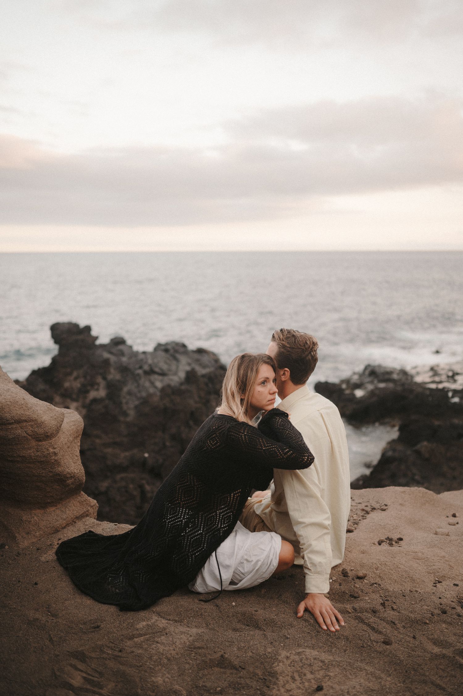 Young couple sitting on a cliff overlooking the ocean, sharing a romantic moment during a photoshoot, Tenerife