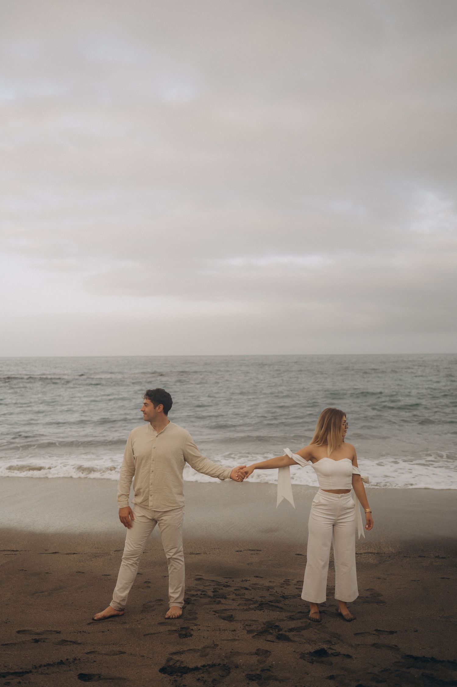 Couple holding hands on Playa del Duque beach in Tenerife with the ocean in the background, enjoying a romantic seaside moment.