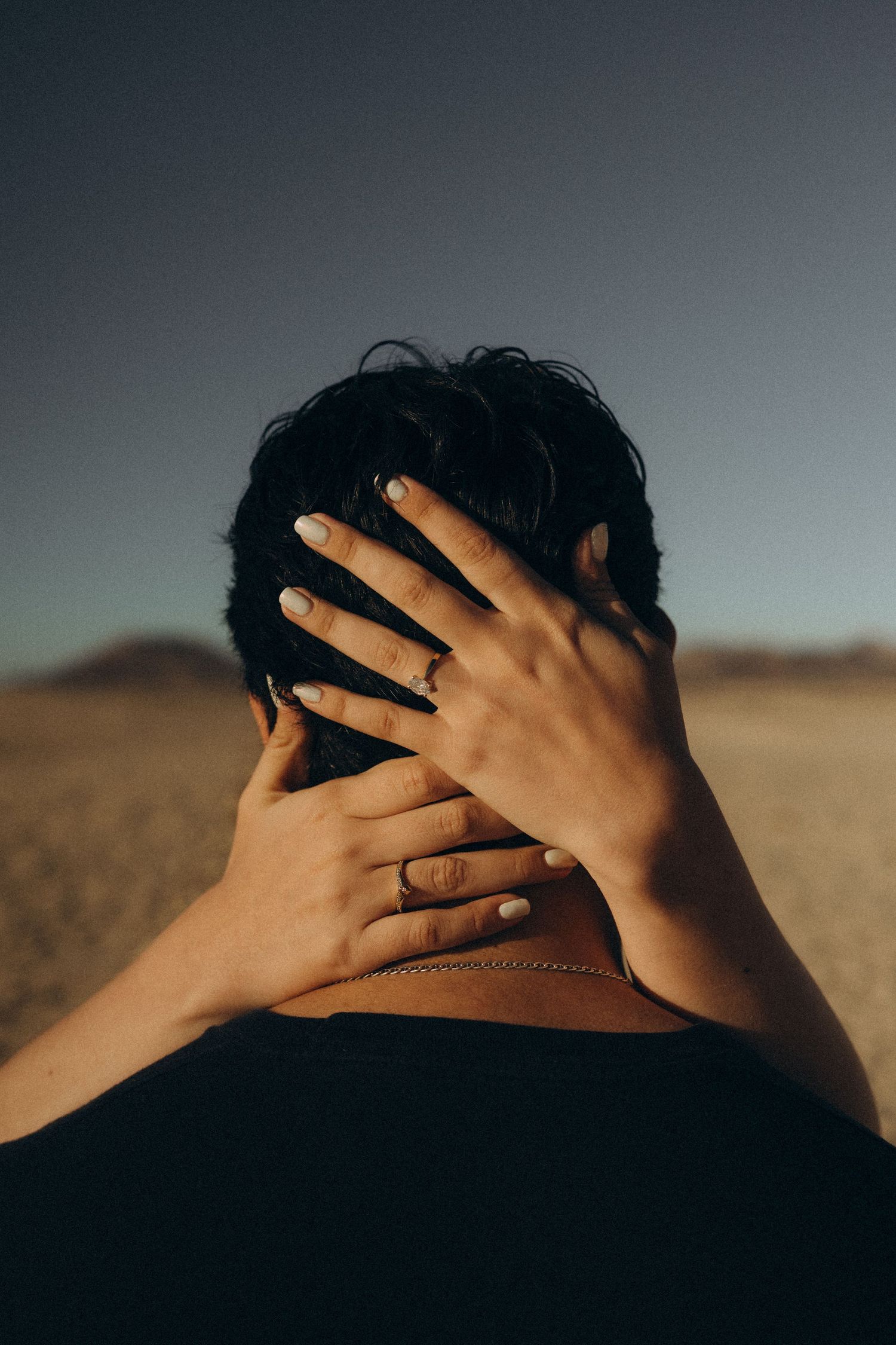 Woman with gold ring cradles mans dark hair with both hands against desert landscape background.
