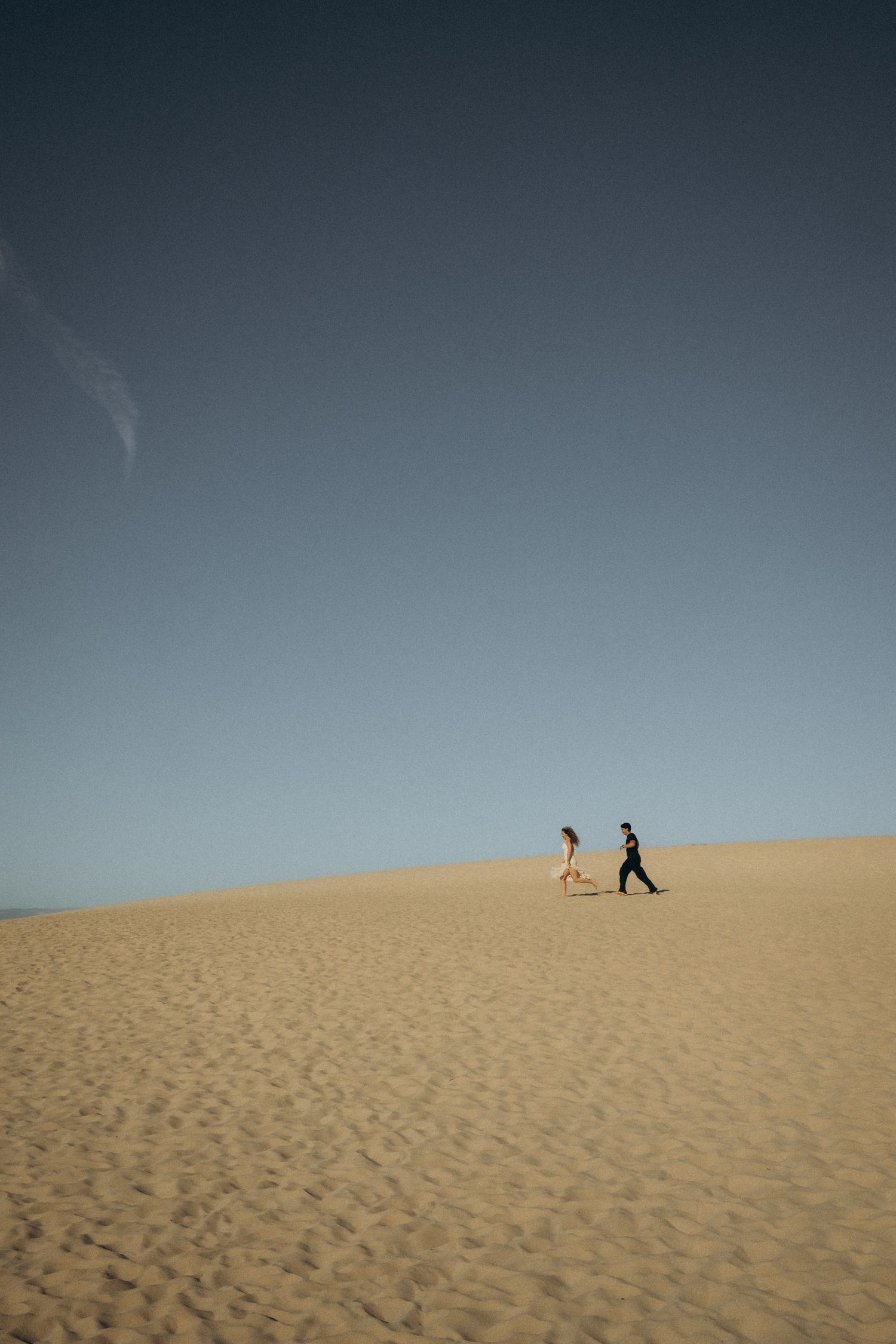Two people walking on a vast sandy desert dune under a clear blue sky.