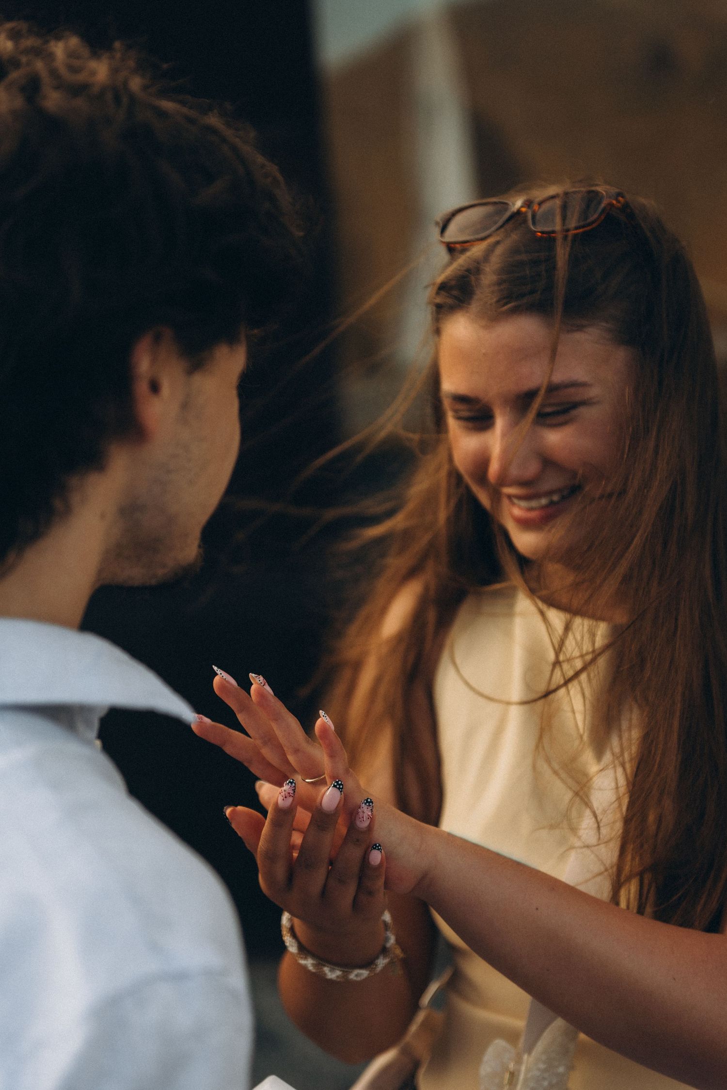 A smiling woman with sunglasses on her head holds hands with a man in a white shirt in a romantic moment.