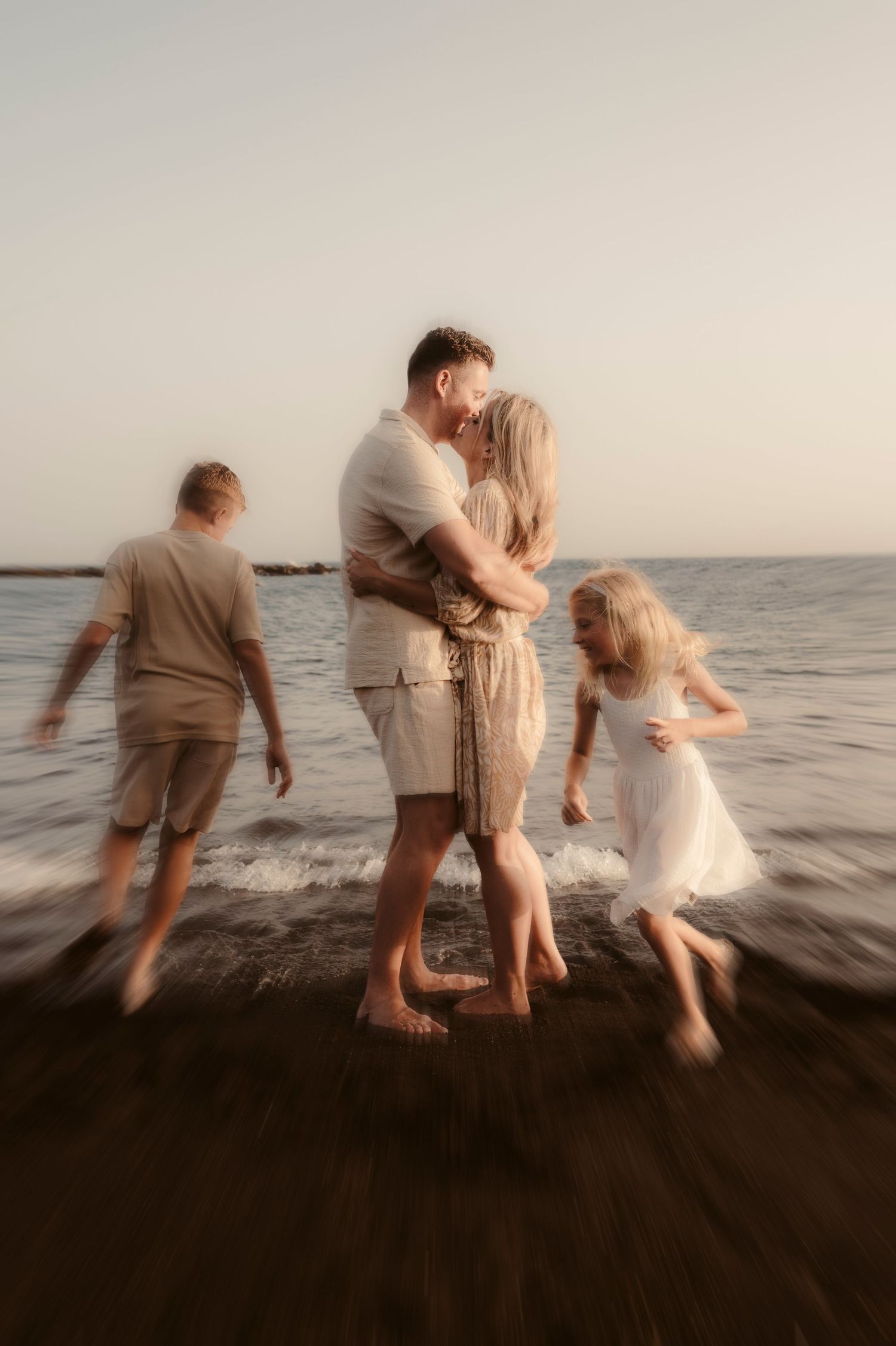 Parents holding each other while kids play nearby on beach at sunset.