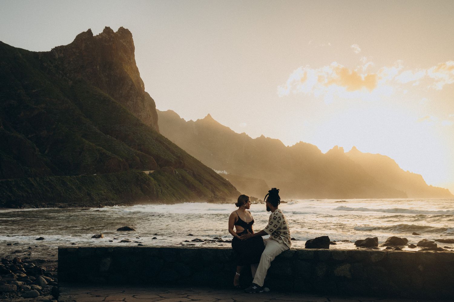 Couple sitting on a stone wall at sunset with dramatic mountain coastline and golden light in the background.