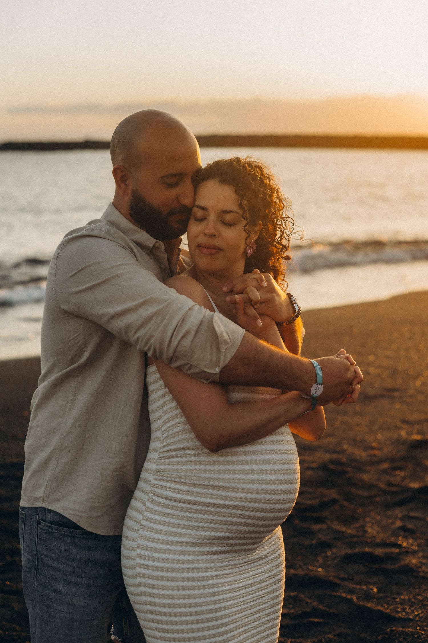 A couple embraces tenderly on a beach at sunset, the woman visibly pregnant, both sharing a peaceful romantic moment.