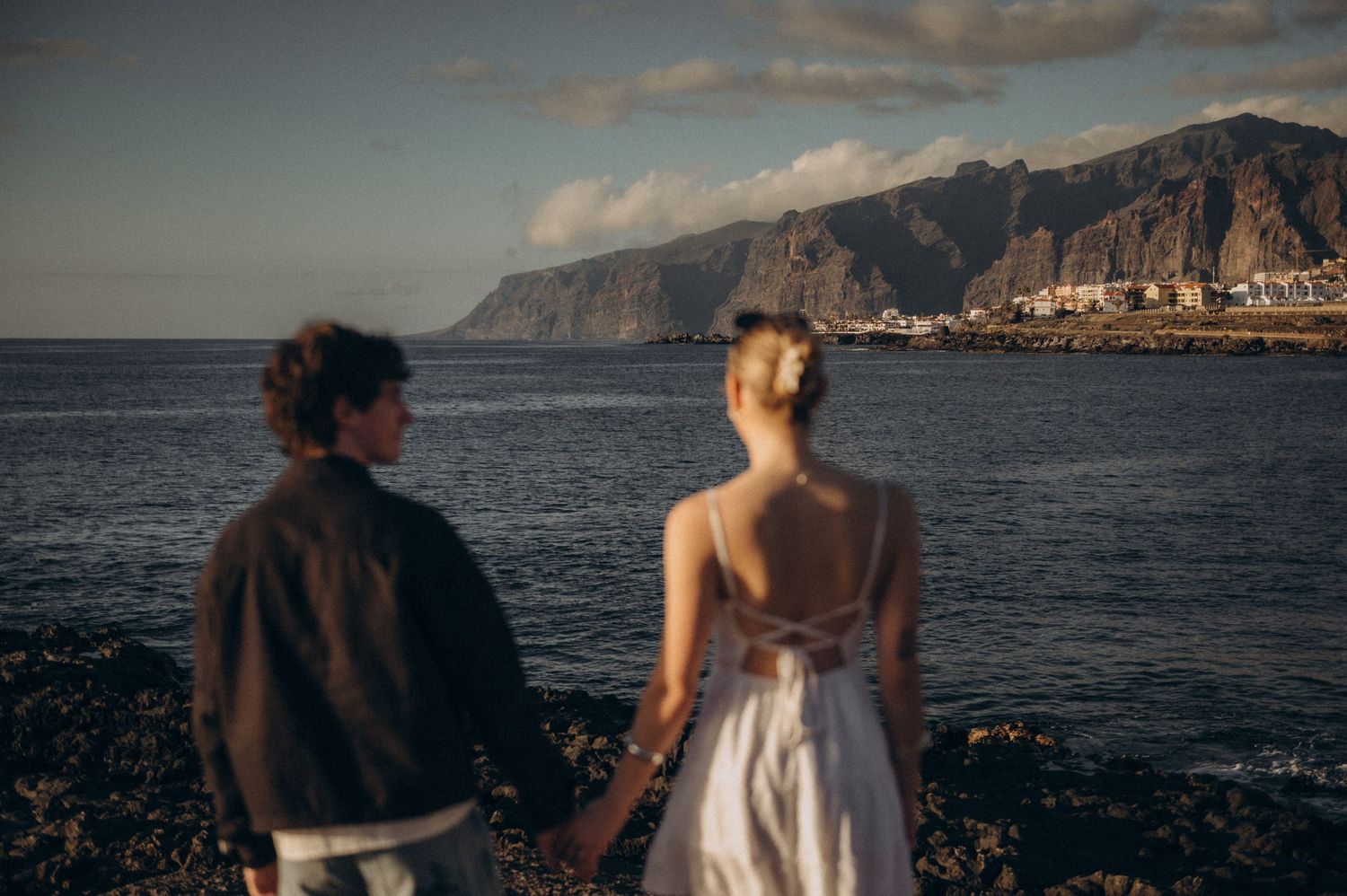 Couple holding hands facing dramatic coastal cliffs and ocean at golden hour, woman in elegant backless white dress.