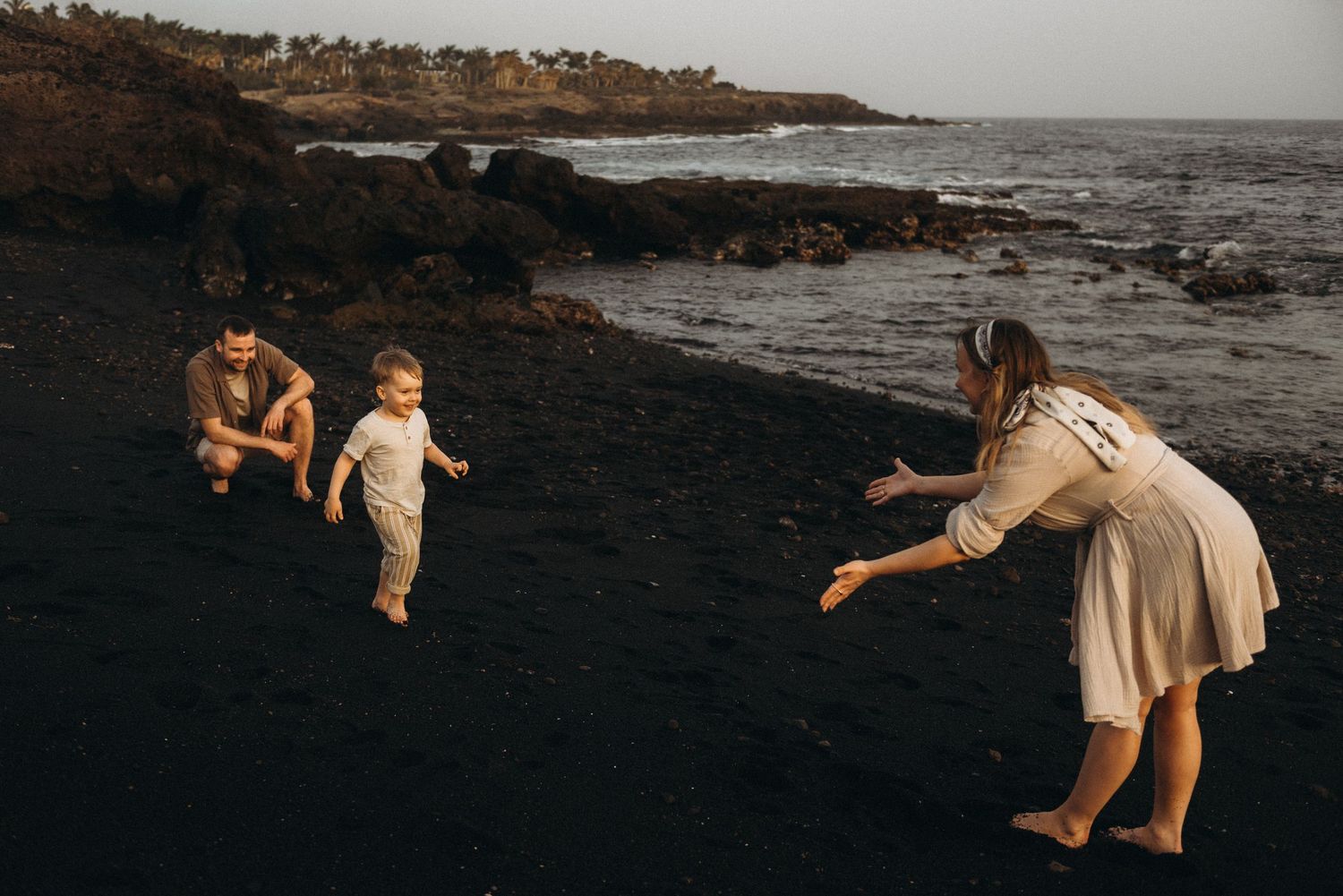 Family playing on a black sand beach, parents reaching for toddler near rocky shoreline at dusk.