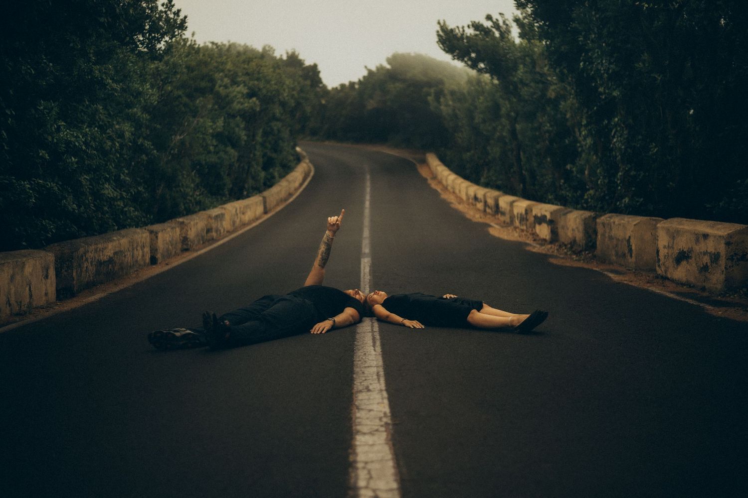 Two people lying on a misty road surrounded by lush green trees, arms outstretched on the center line.