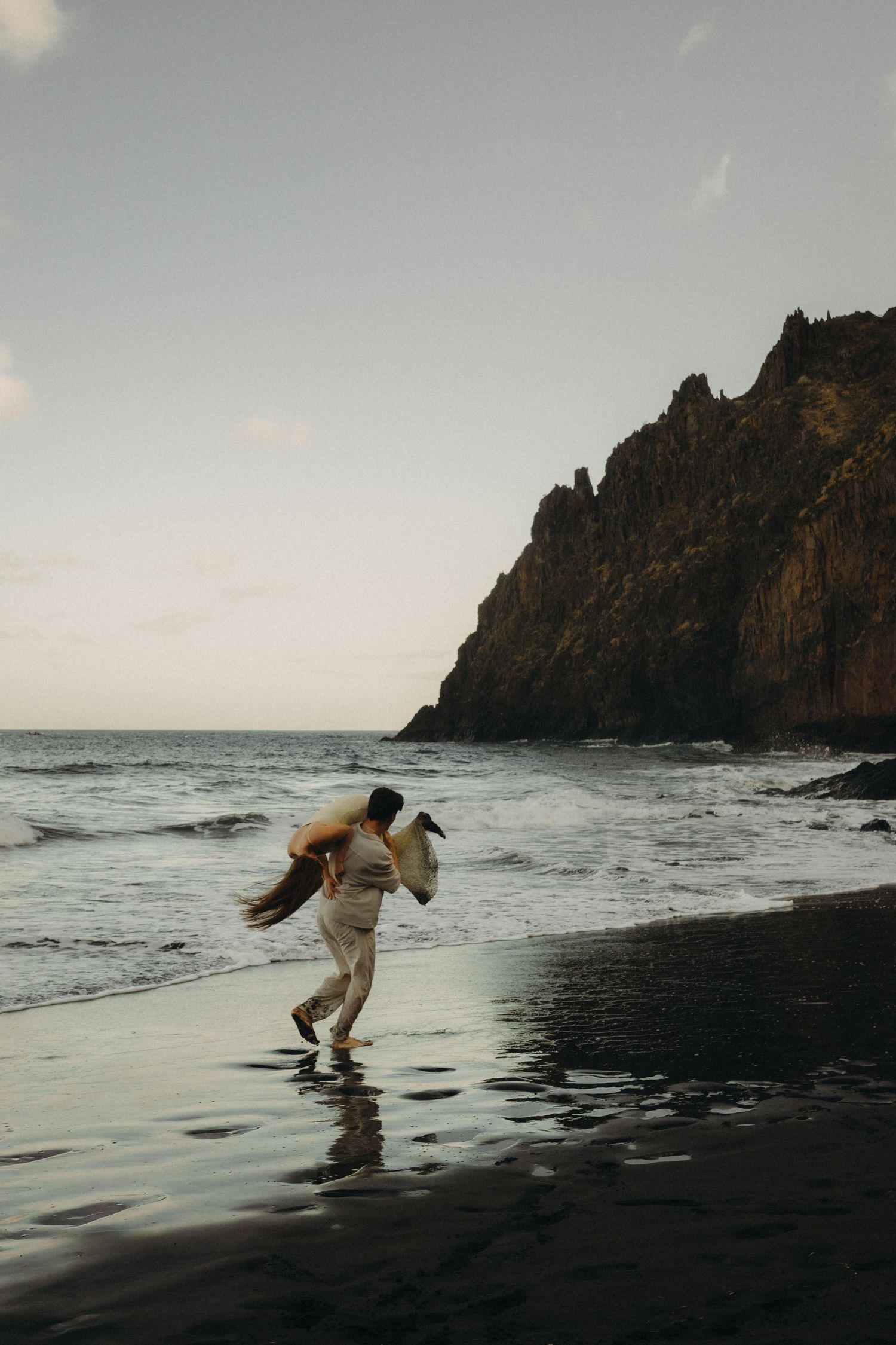 Couple running on a black sand beach at dusk with dramatic rocky cliffs and ocean waves in the background.