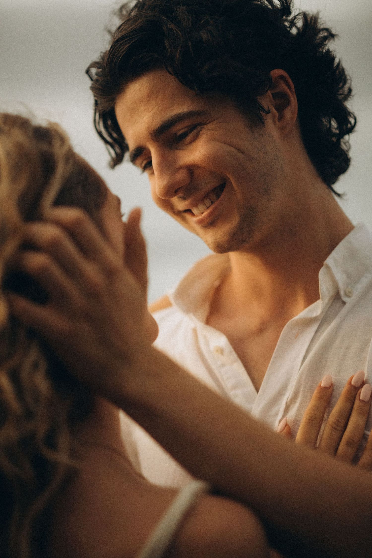 A smiling man with curly hair in a white shirt gazes tenderly at a woman, holding her face in a romantic moment.