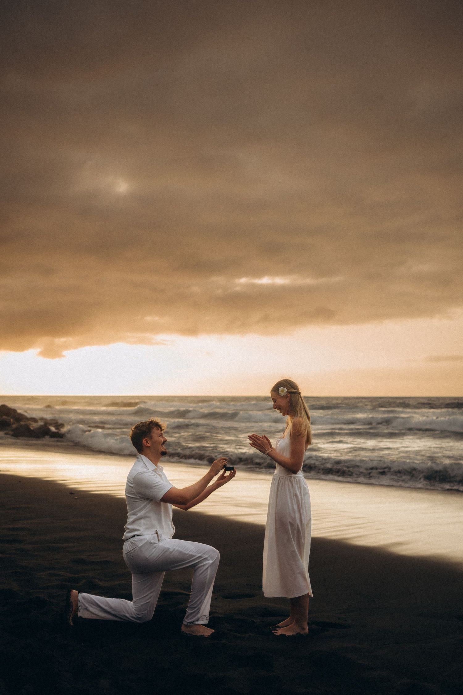 Man proposing to woman on black sand beach at sunset, both dressed in white, with dramatic cloudy sky.