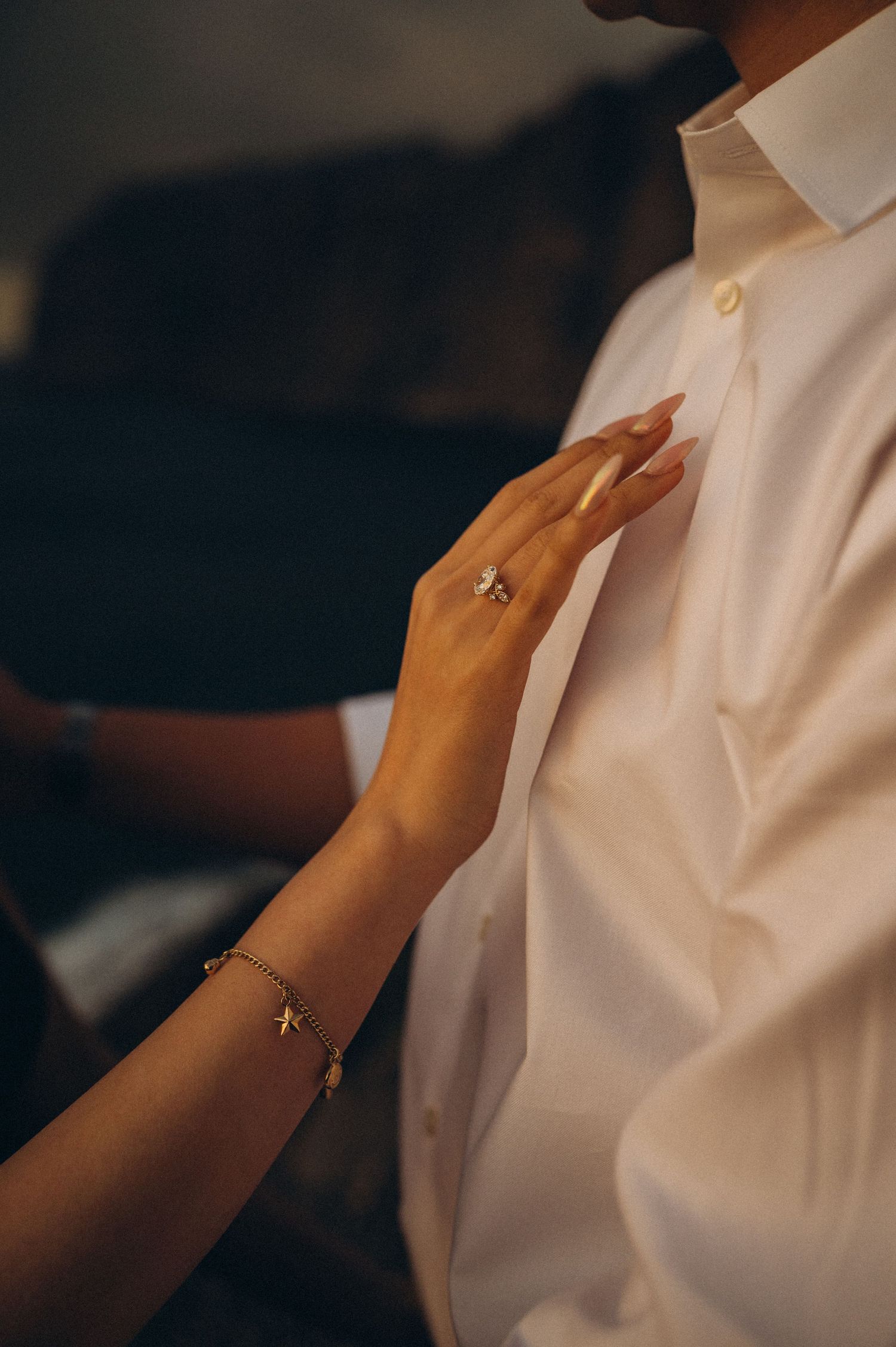 A woman's hand with a diamond ring and gold bracelet rests on a man's white dress shirt.