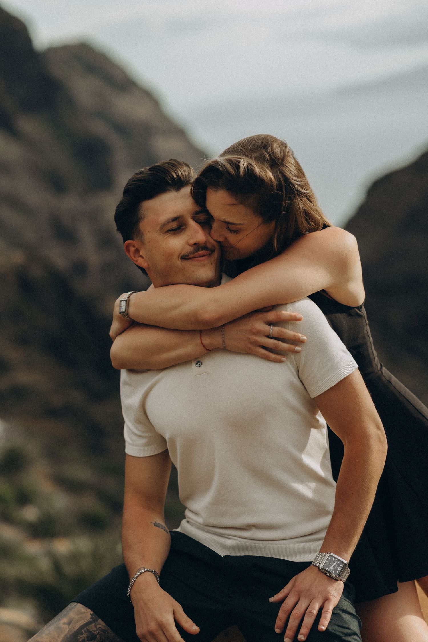 A couple embraces outdoors, woman hugging man from behind near rocky mountains, romantic lifestyle photography.