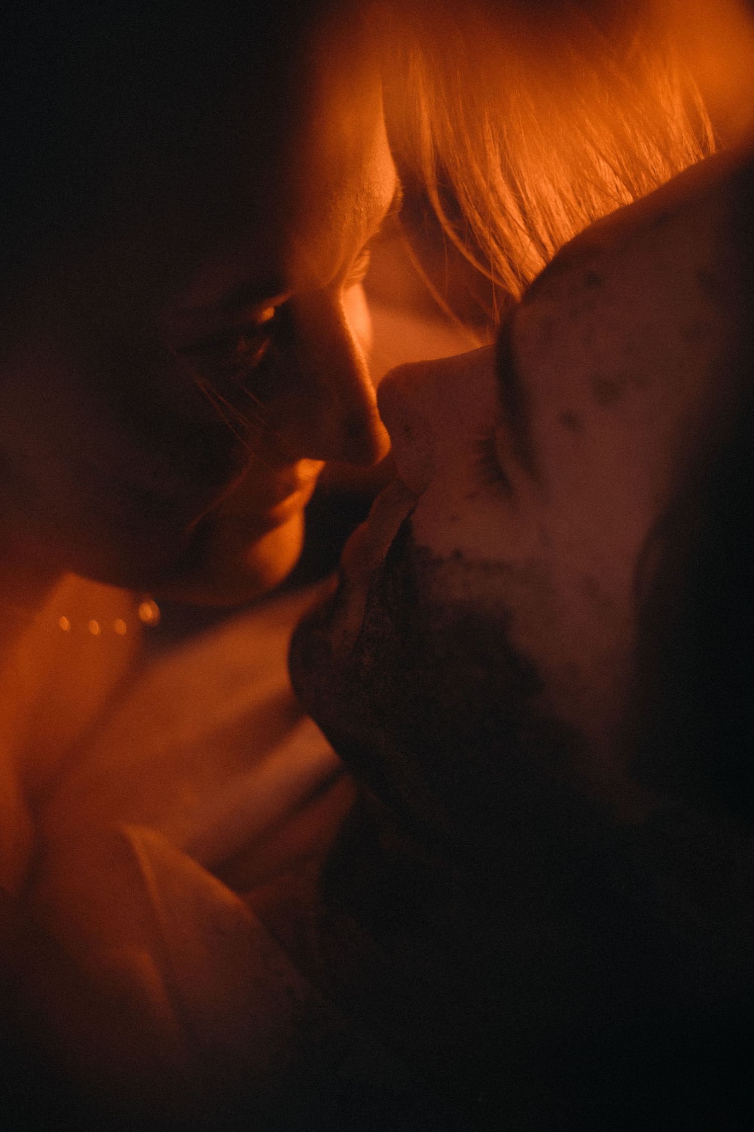 Two people share an intimate moment bathed in warm orange candlelight glow in a romantic close-up portrait.