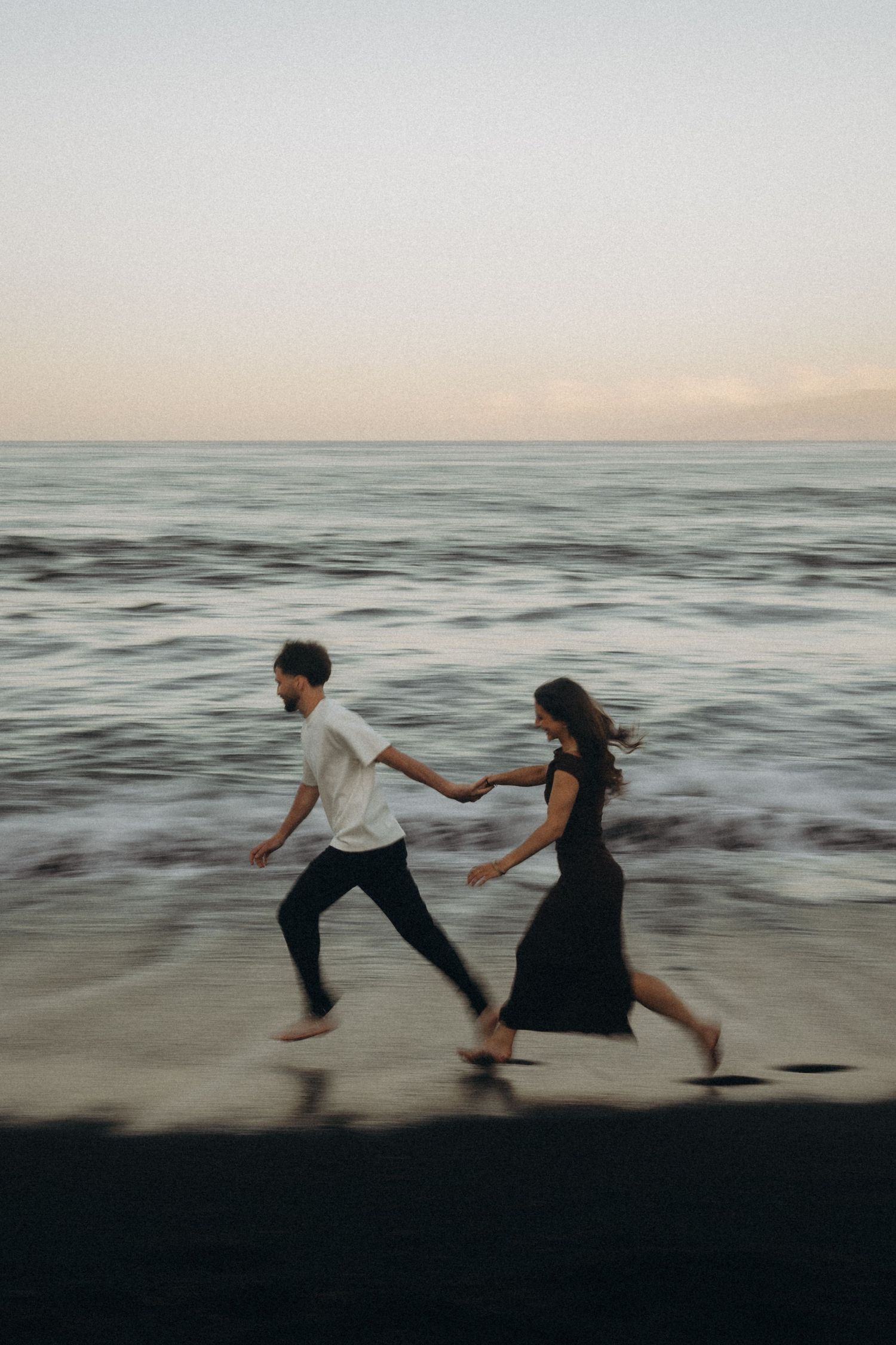 A couple runs hand-in-hand along a beach at dusk, with ocean waves in the background, captured with motion blur.