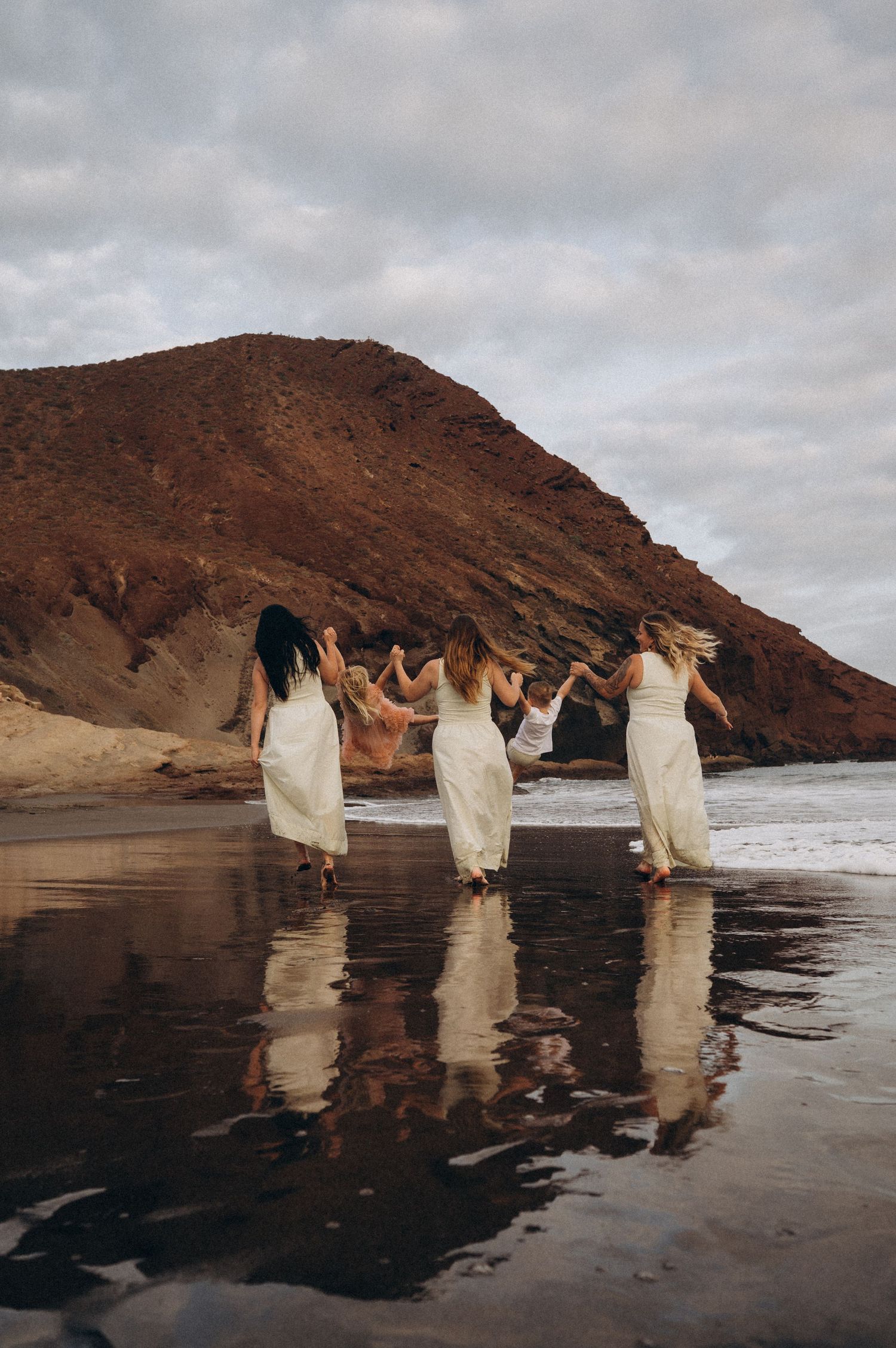 Three women walking side by side lifting toddlers, reflections in sand, Montana Roja Tenerife