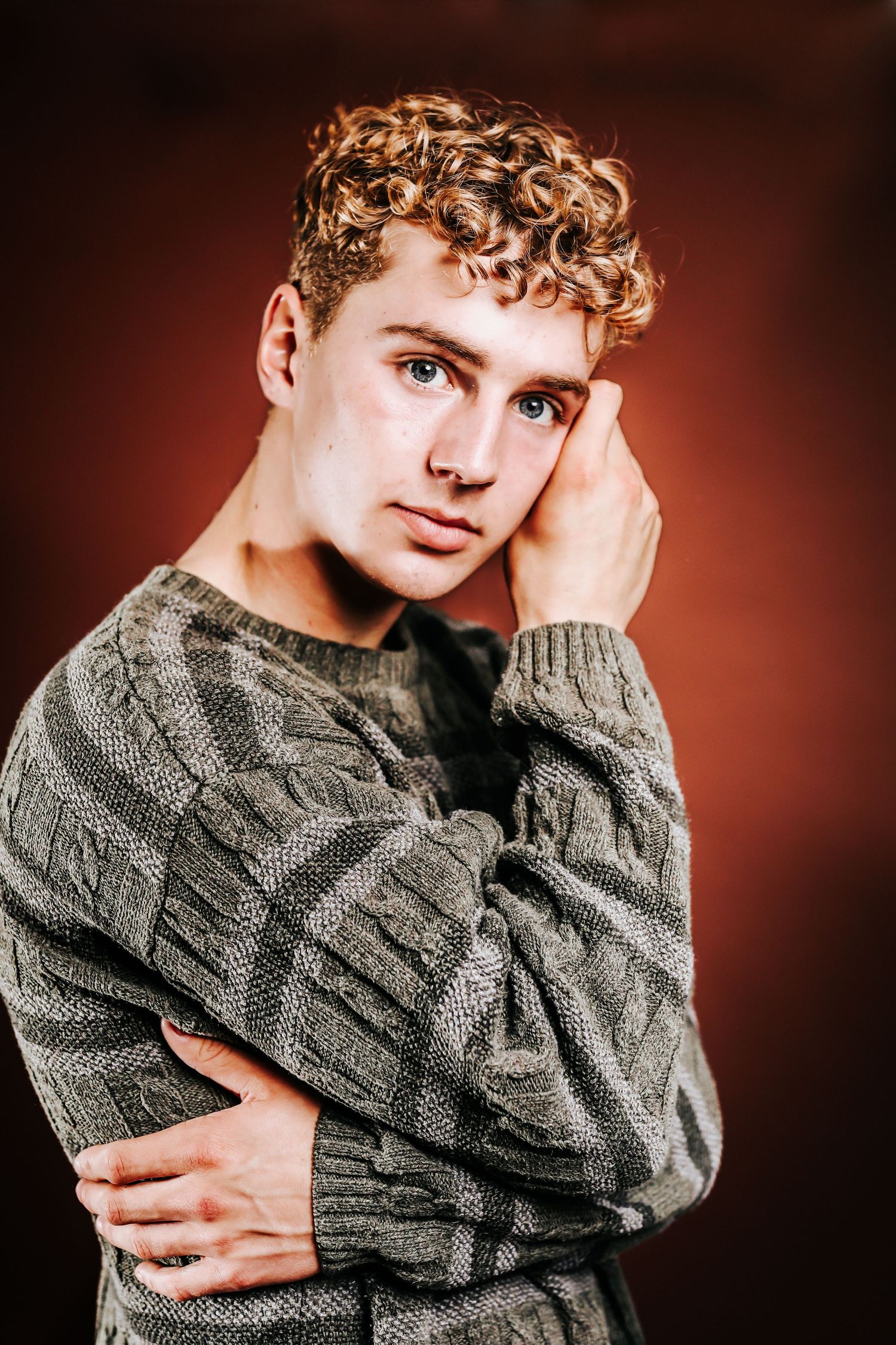 Subject wearing gray cable knit sweater poses against warm red backdrop in studio portrait series.