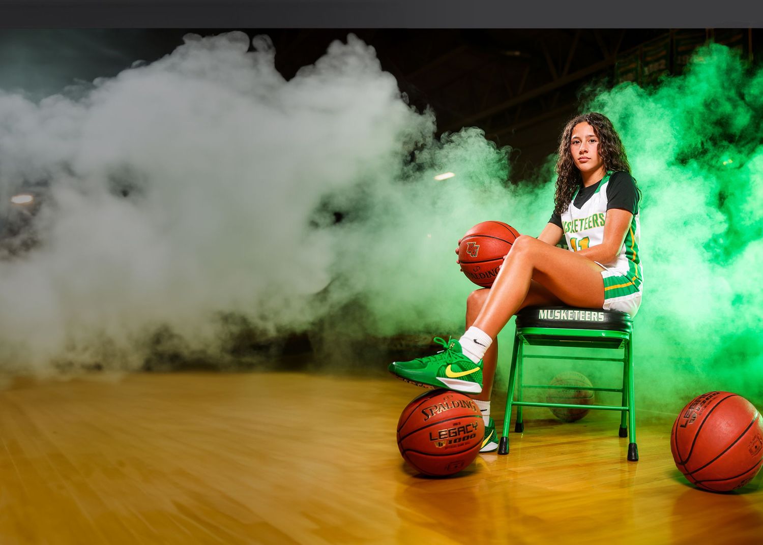 Basketball player poses on chair surrounded by green and white smoke effects on wooden court with basketballs.