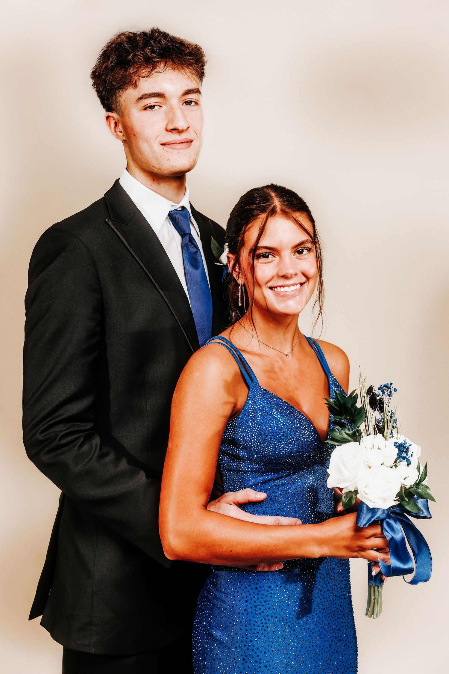 A couple poses in formal attire at a school dance, with the woman wearing a blue gown and holding a white bouquet.