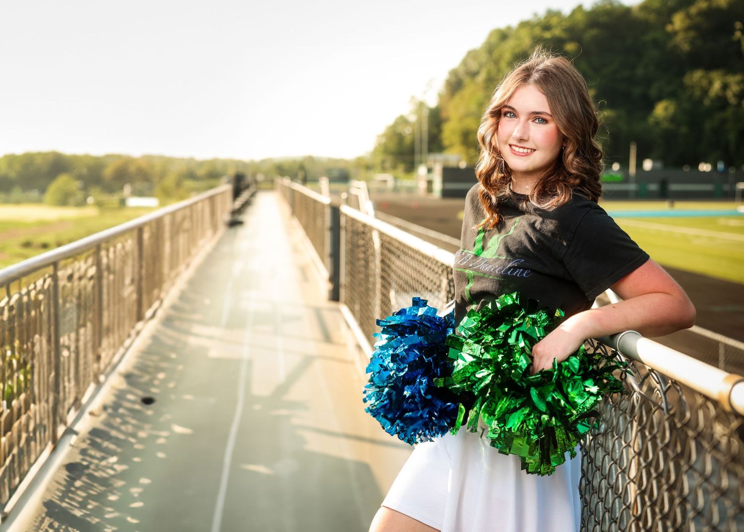 A cheerleader in a white uniform holds blue and green pom poms while posing on a wooden bridge during sunset.