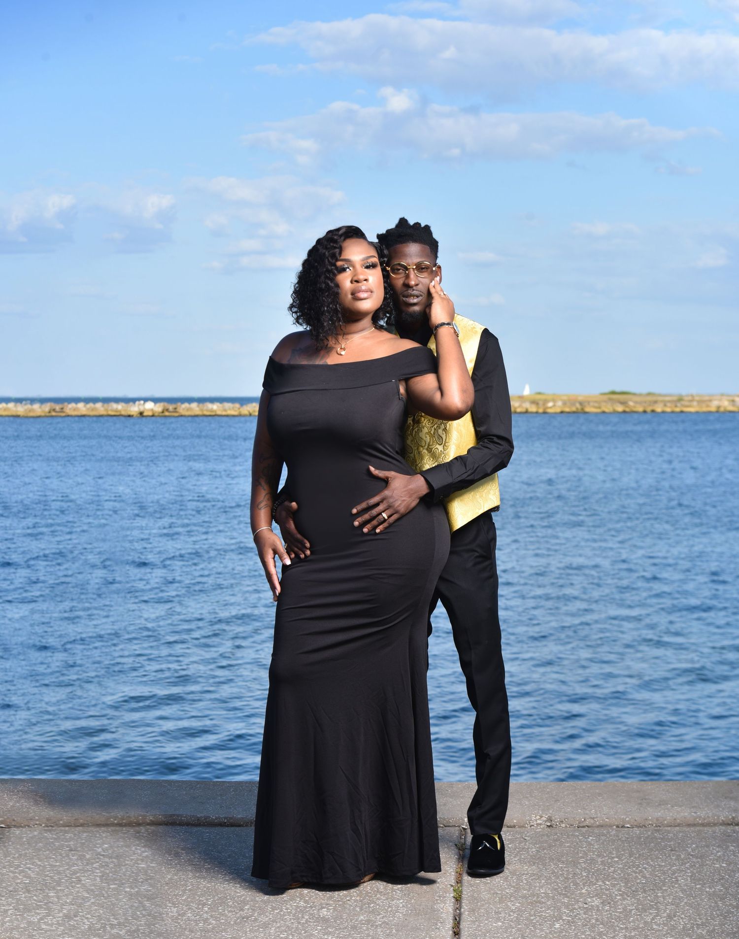 Couple posing by waterfront, woman in black off-shoulder gown, man in black suit with gold vest.