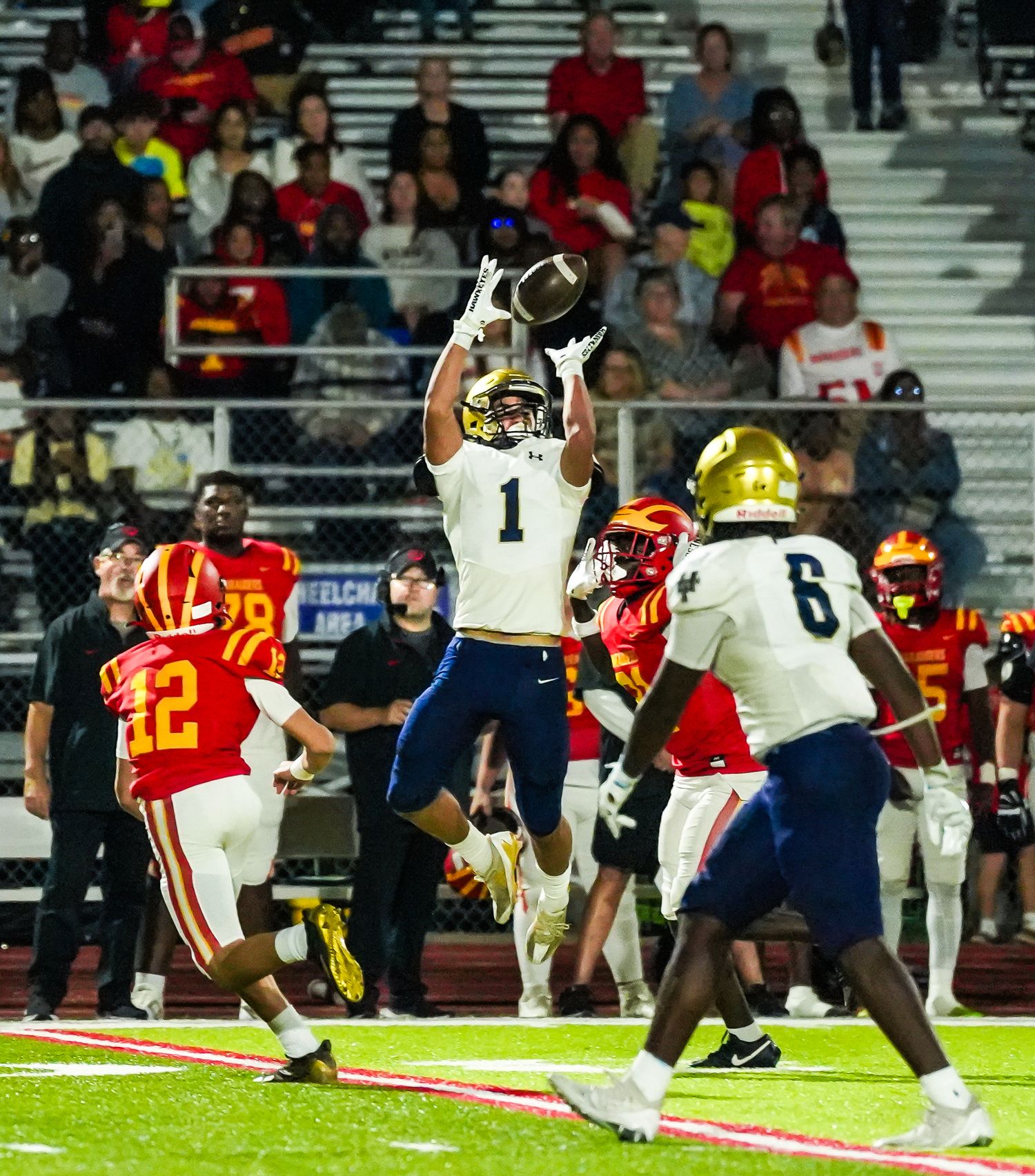 Football player #1 in navy uniform leaps to catch a pass during a high school game against red-uniformed opponents.