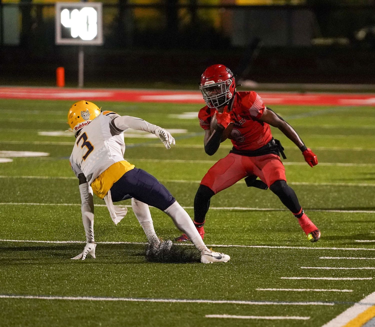 Football player in red uniform makes a move past a defender in yellow during a night game on a green turf field.