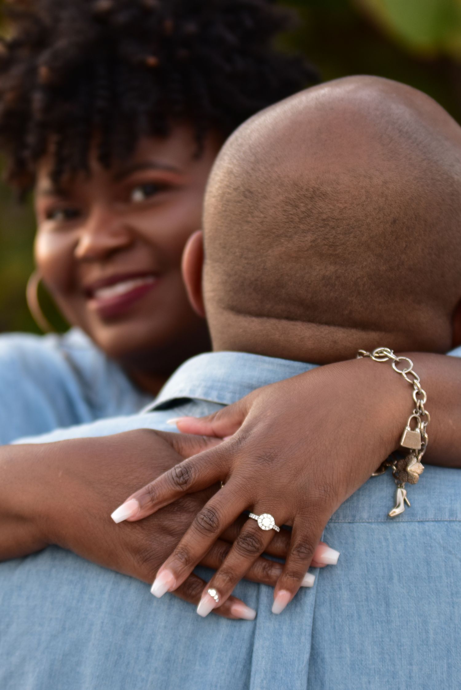 A joyful woman hugs a man while showing off her engagement ring and gold charm bracelet, both wearing denim.