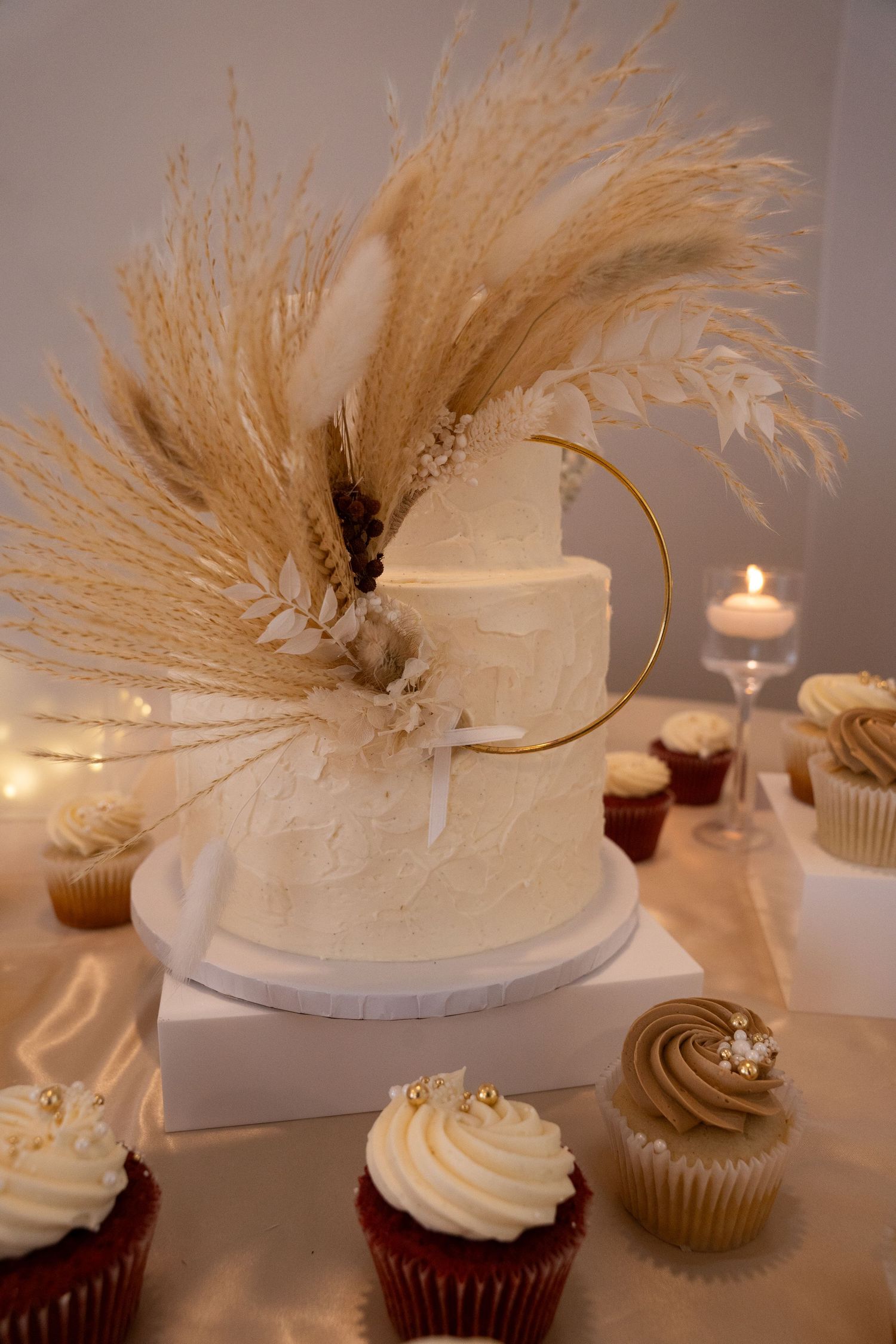 Elegant white wedding cake decorated with dried pampas grass and gold hoop, surrounded by cupcakes in Saint Petersburg Florida.