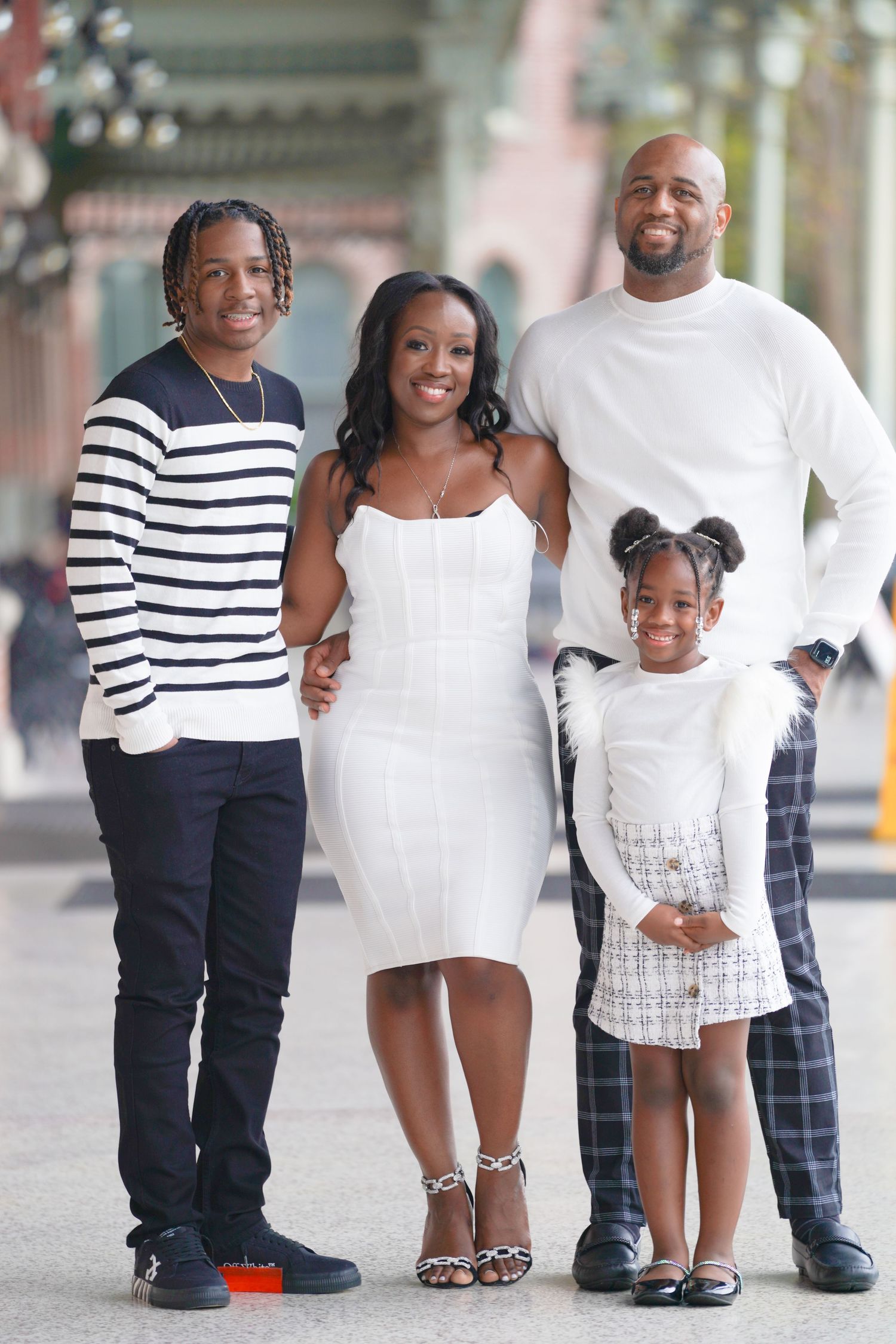 A joyful family of four poses together outdoors, dressed in white and casual attire, smiling warmly for a portrait.