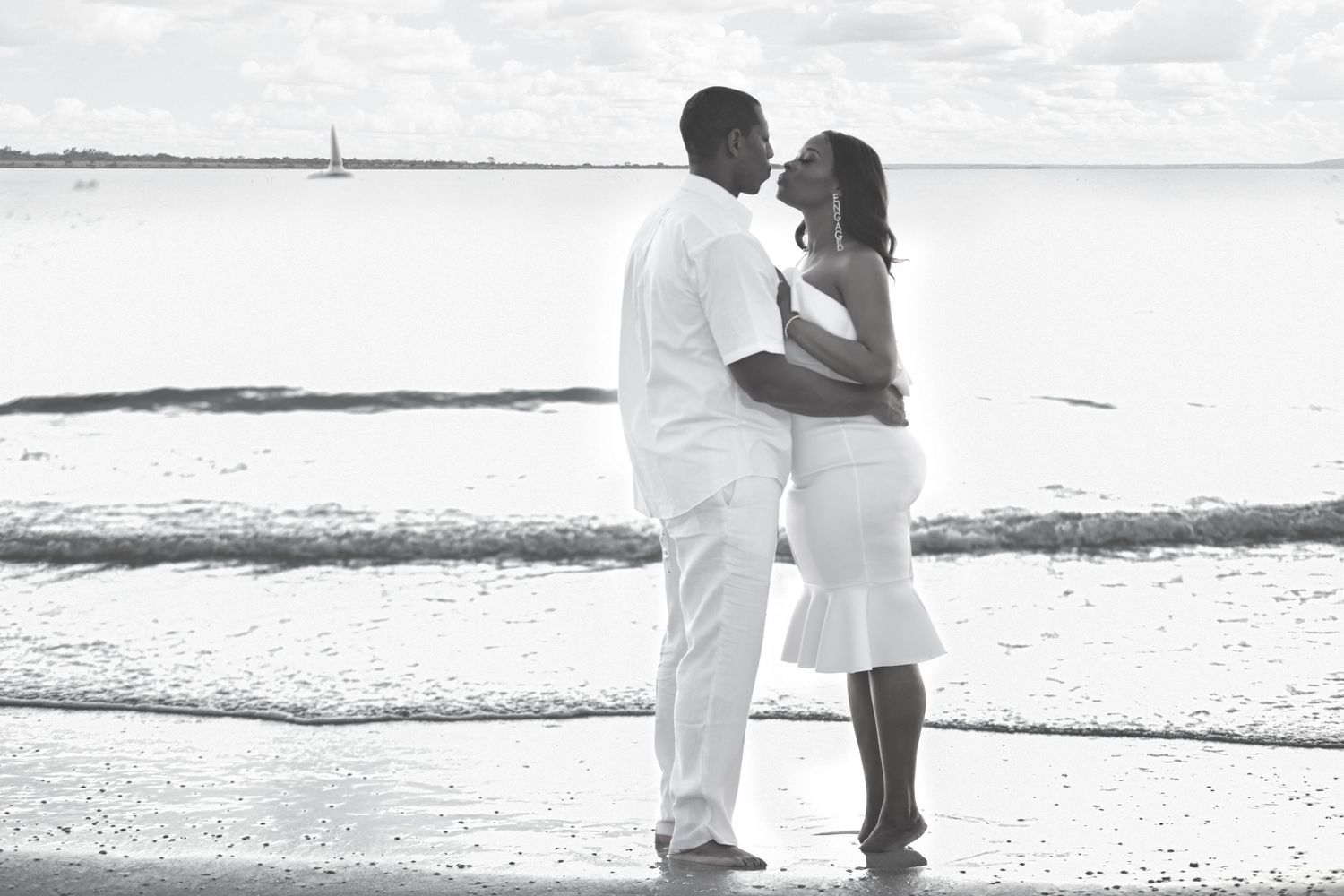 Black and white photo of a couple kissing on a beach, woman in white dress showing baby bump, man in white outfit.