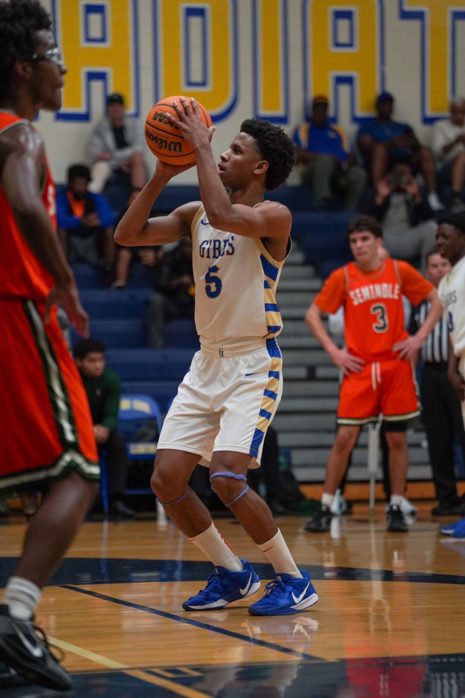 High school basketball player #5 in blue and gold uniform holds ball during indoor game against Seminole team.