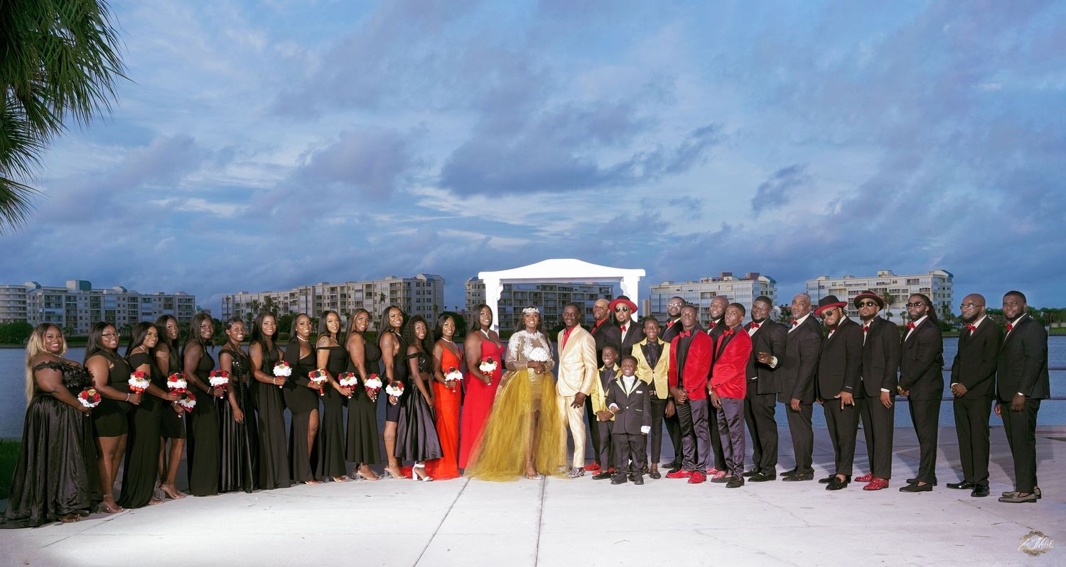 Large wedding party lined up on white sand beach wearing formal attire.