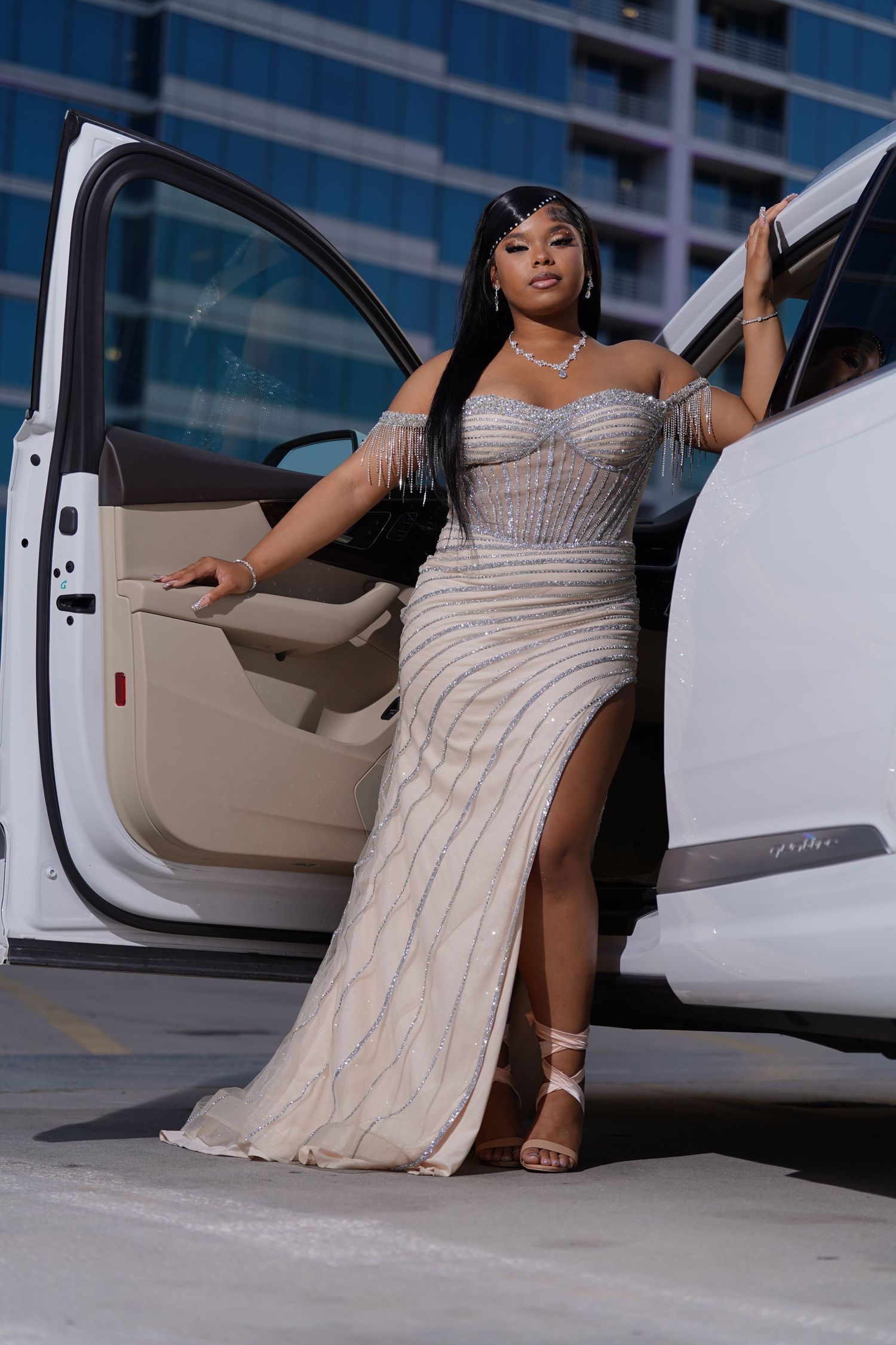 Woman in elegant beaded gown with slit poses beside white luxury car on rooftop parking deck.