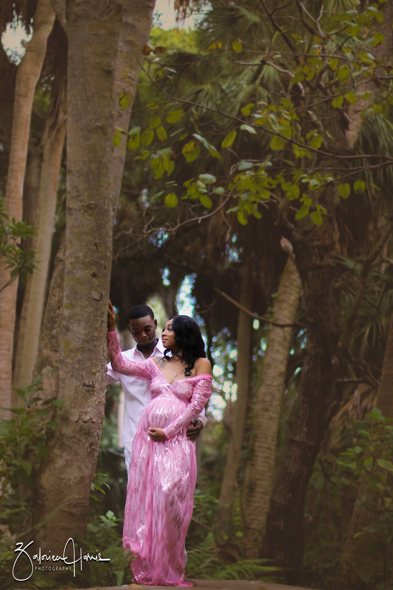 A person in a flowing pink gown poses elegantly among tall trees in a dreamy forest setting.