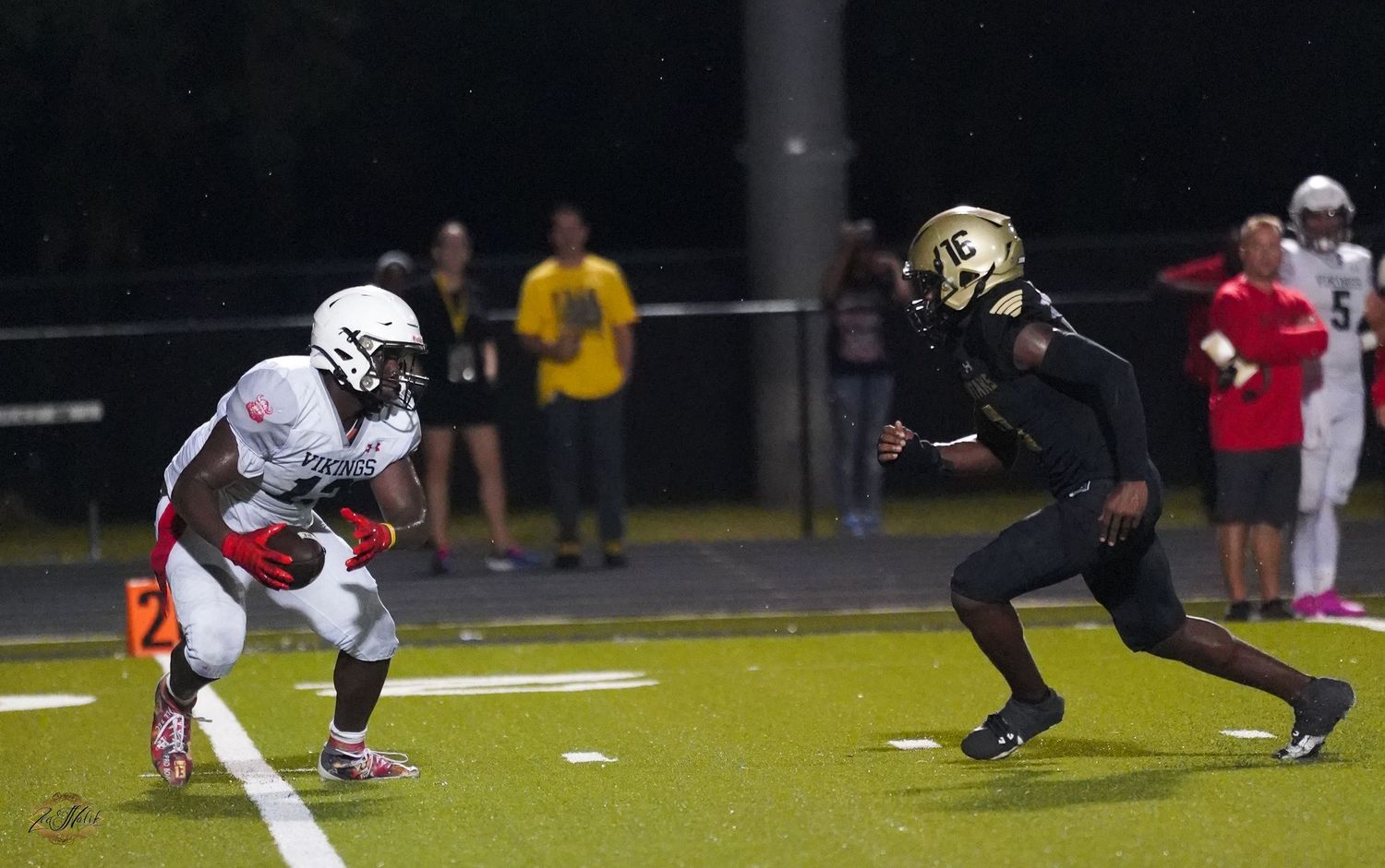 Two football players compete on a night game field, one in red and white uniform carrying the ball, pursued by a defender.