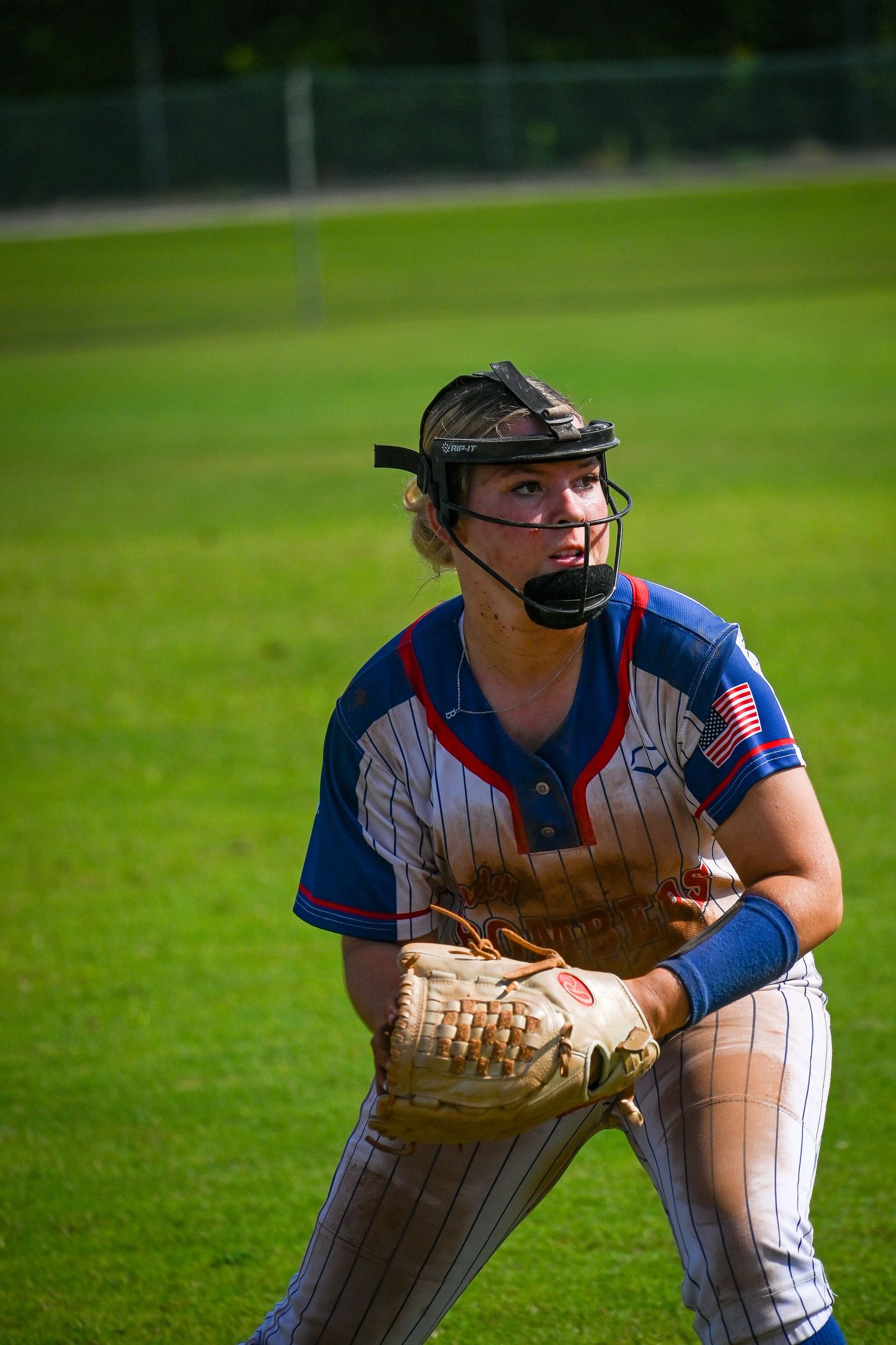 Female softball player in blue jersey with face guard holds glove ready on green field.