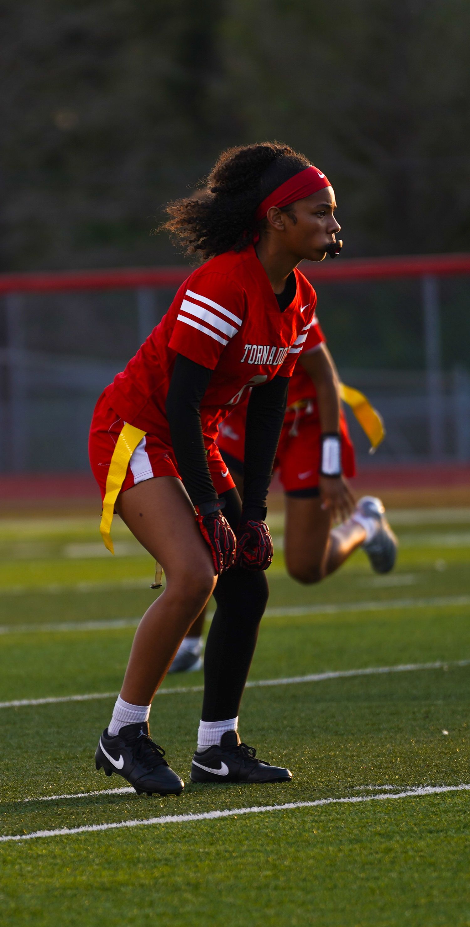 Female athlete in red Tornados jersey plays flag football on green turf field during evening game.