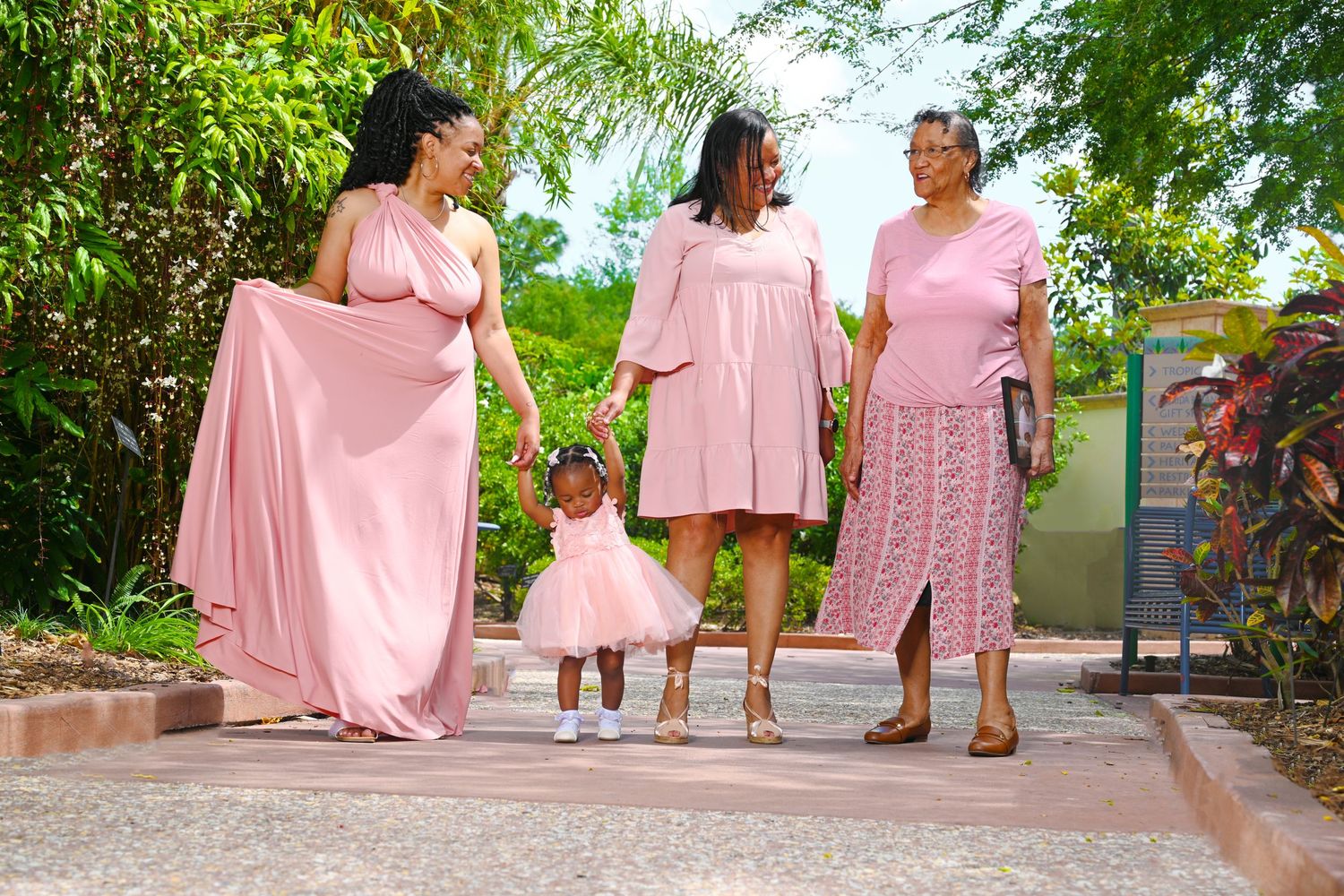 Four generations of women in matching pink outfits walk together outdoors, sharing a joyful family moment.