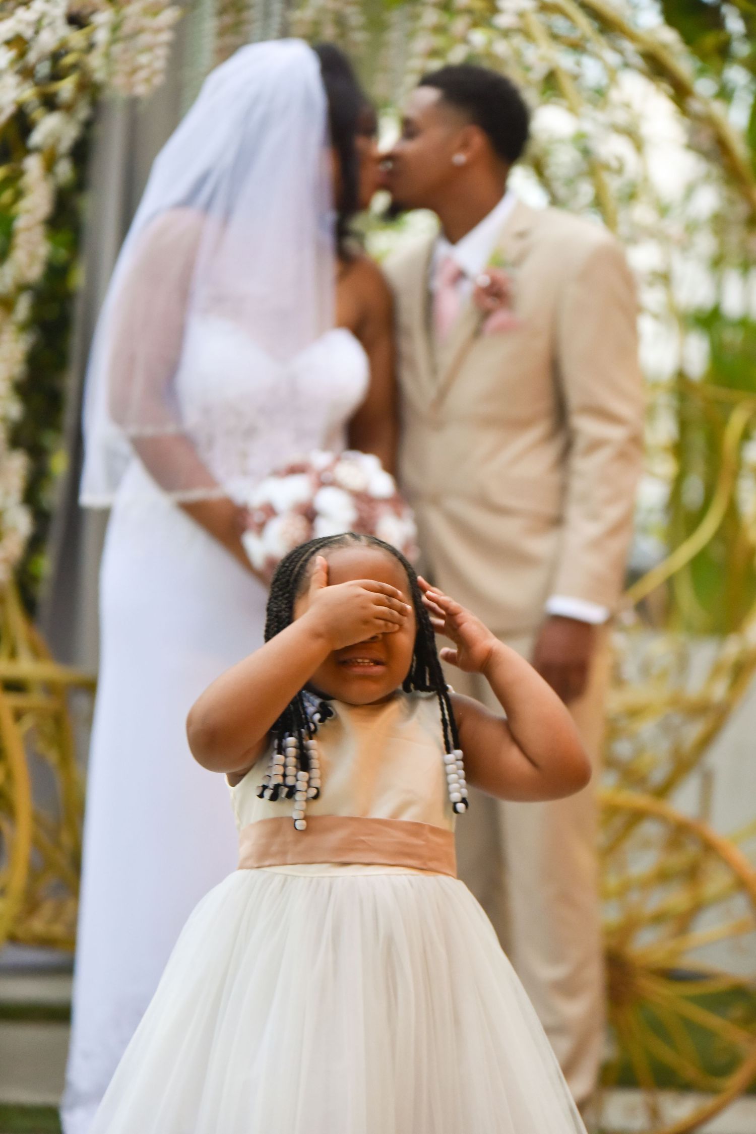 A flower girl covers her eyes as a bride and groom share a kiss at their outdoor wedding ceremony.