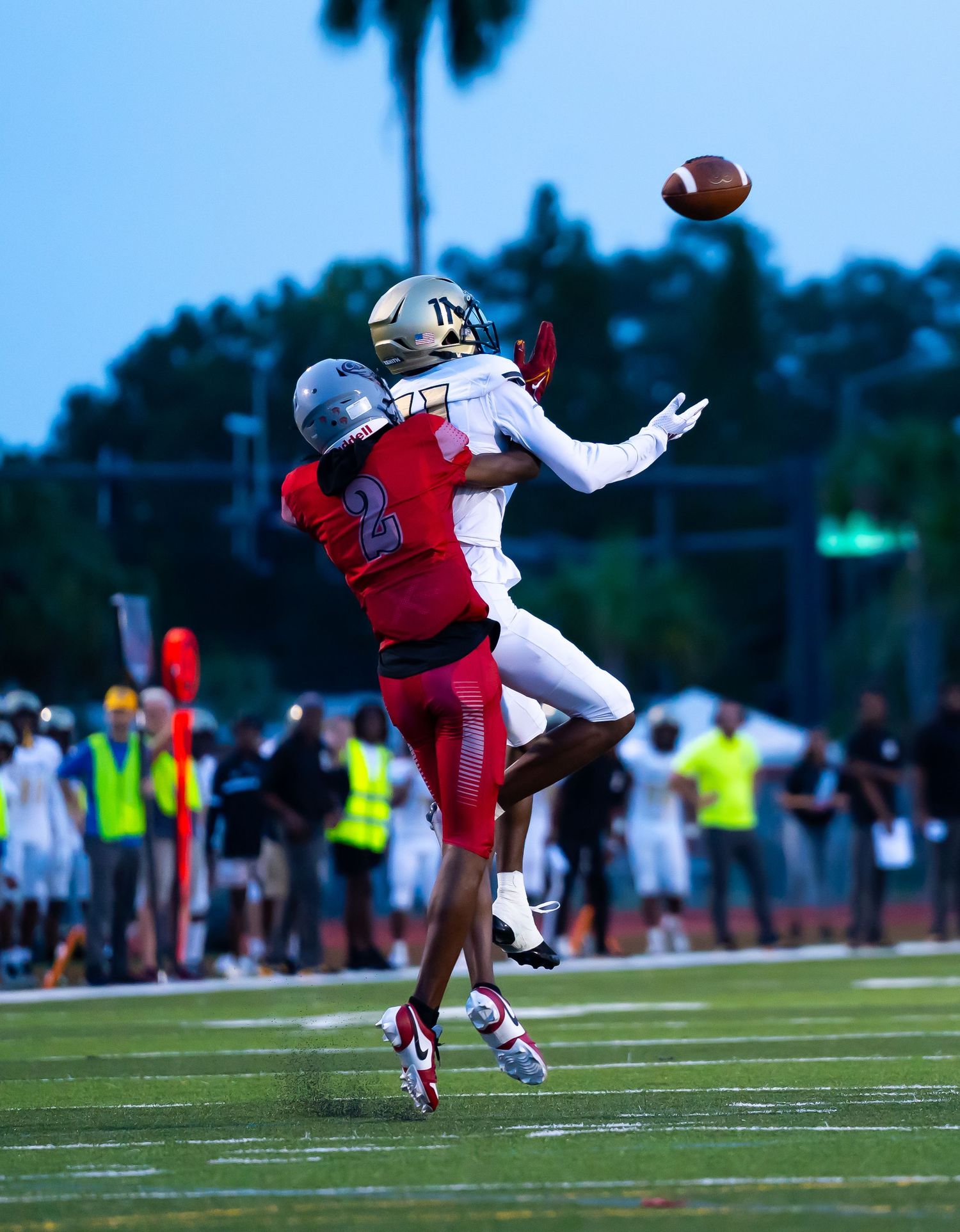 Two football players jump for a contested pass during a high school night game under stadium lights.
