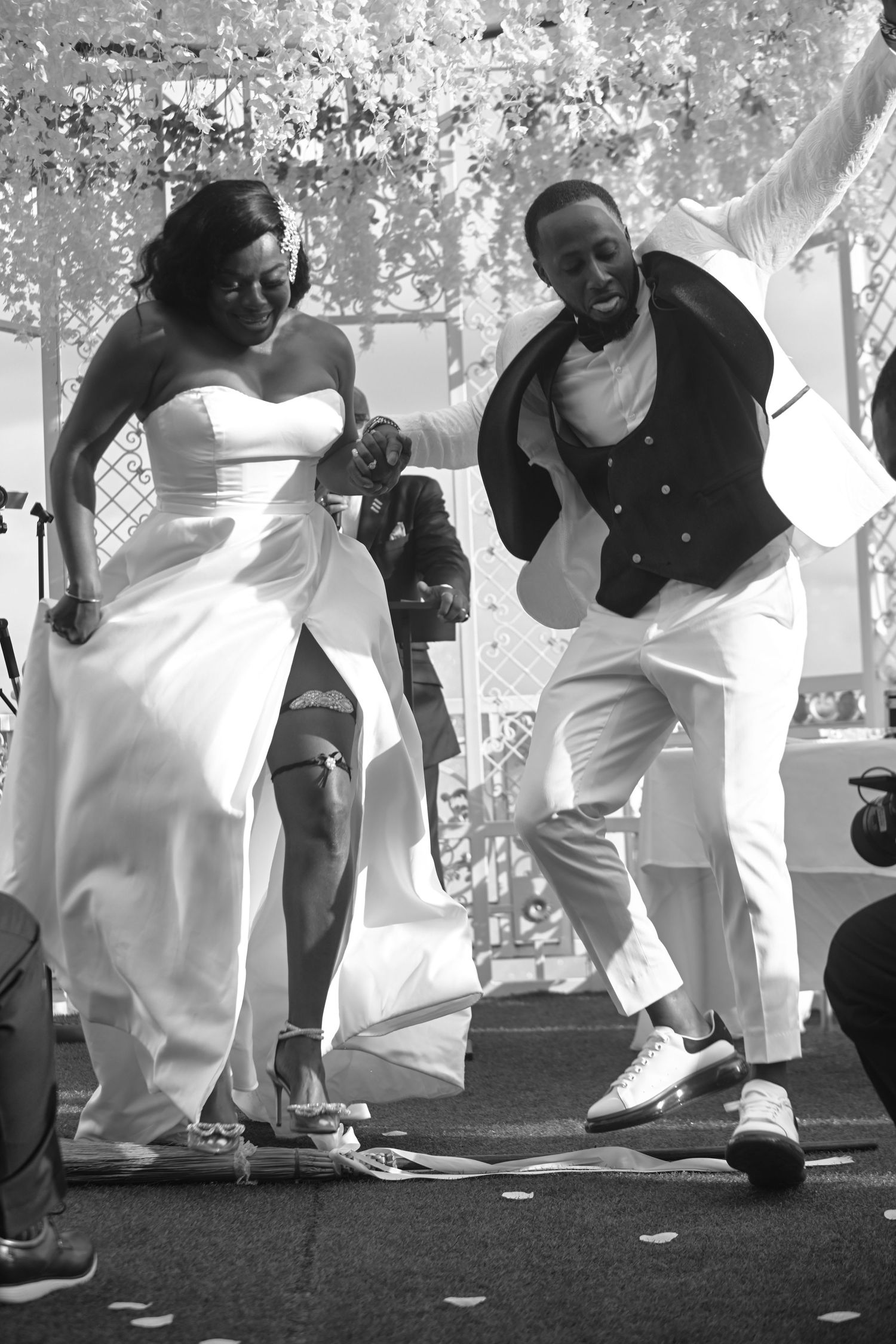 Black and white photo of a joyful bride and groom dancing energetically at their outdoor wedding celebration.