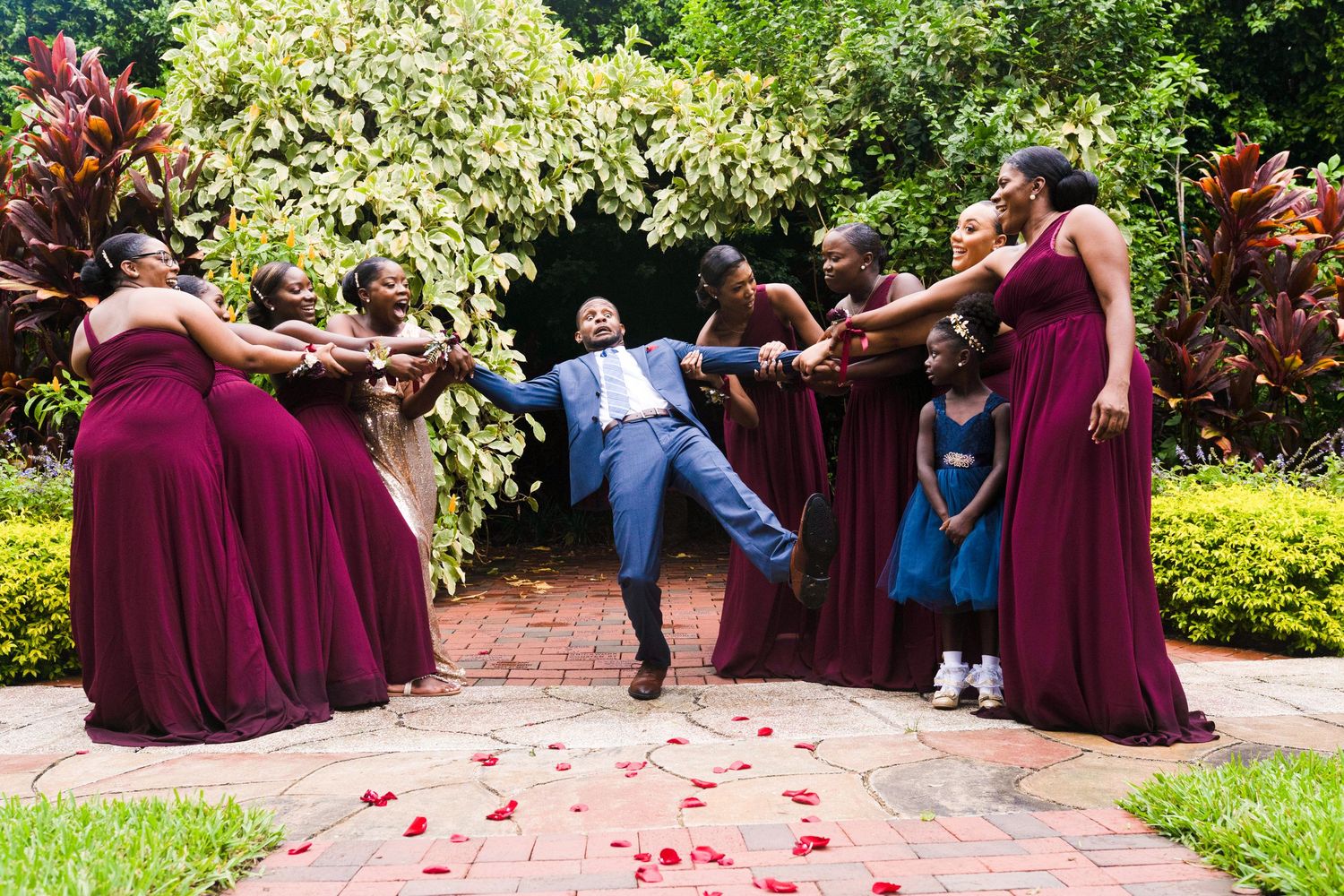 Groom in blue suit lifted by bridesmaids in burgundy gowns on a rose petal-lined path at an outdoor wedding.
