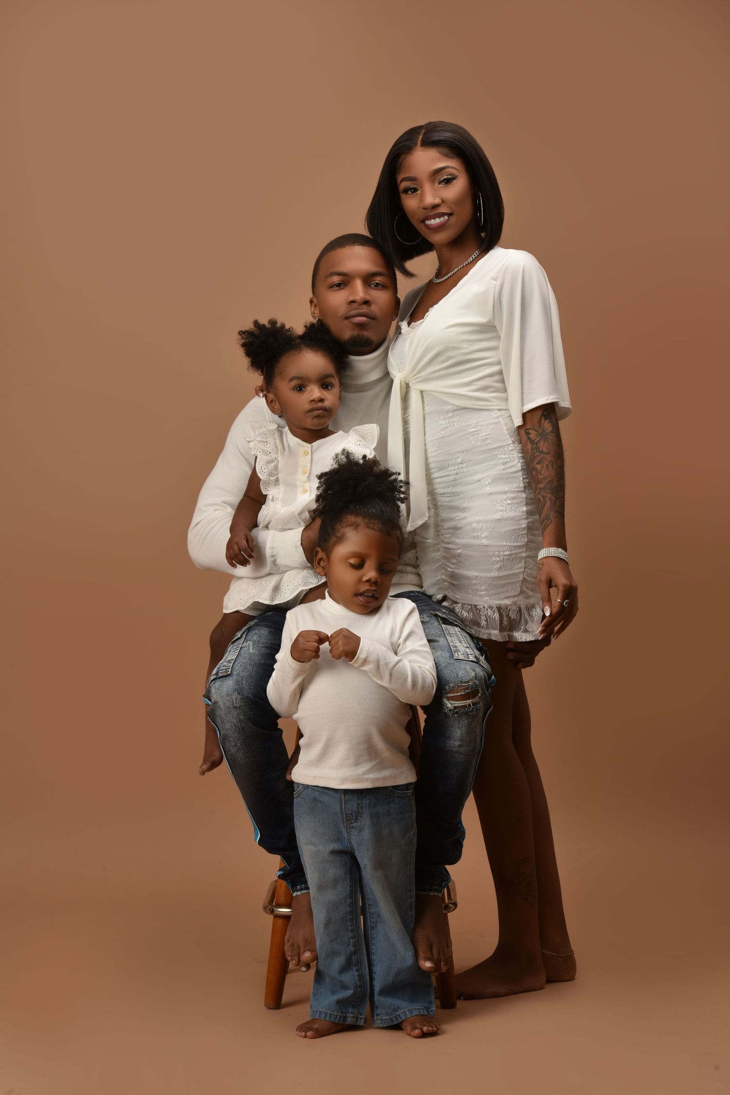A Black family of four poses in a studio, parents in white outfits with two young children in jeans against a brown background.