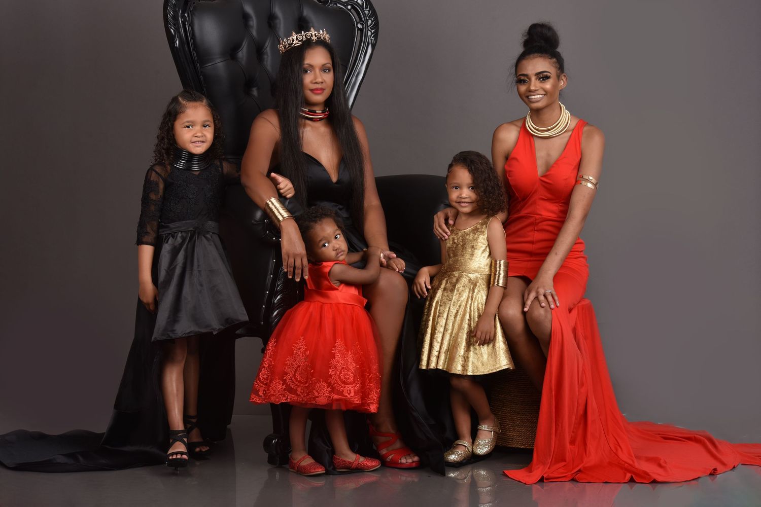 Two women in elegant red and black gowns pose with three young girls in matching formal dresses for a glamorous family portrait.
