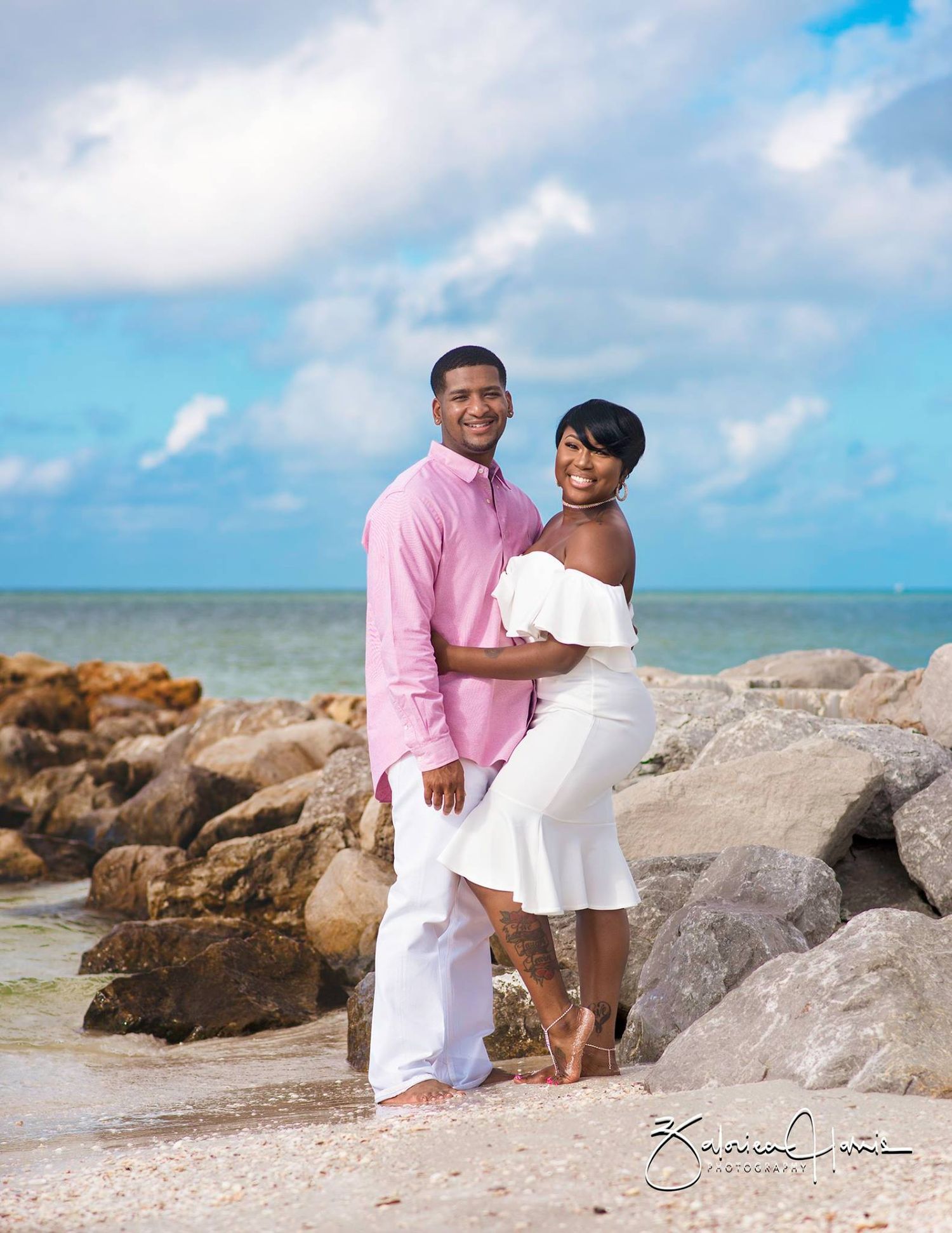 Couple enjoying a romantic moment together on a rocky beach with blue ocean waters and cloudy sky in the background.