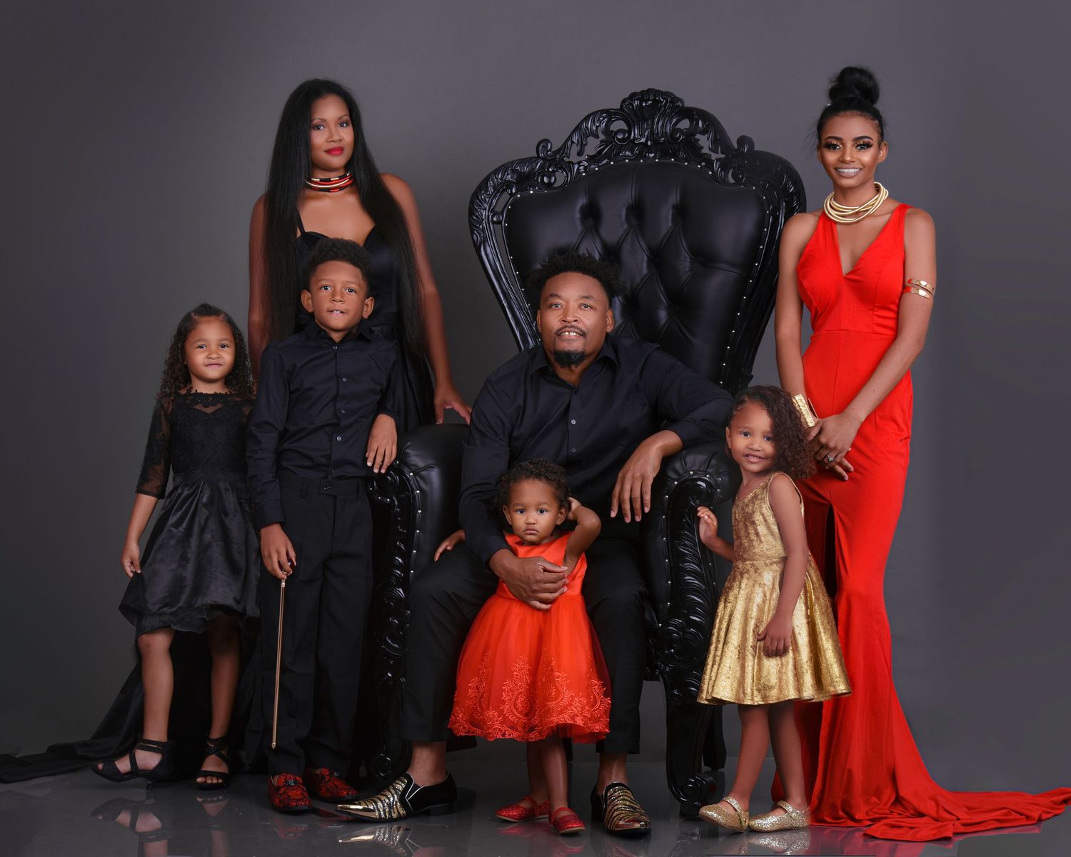 Family portrait with seven members in elegant red, black, and gold outfits posed around a decorative throne chair.