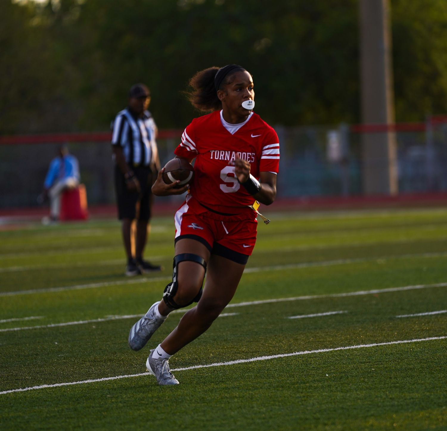 Female athlete in red Tornadoes uniform runs with football on green field during evening game.